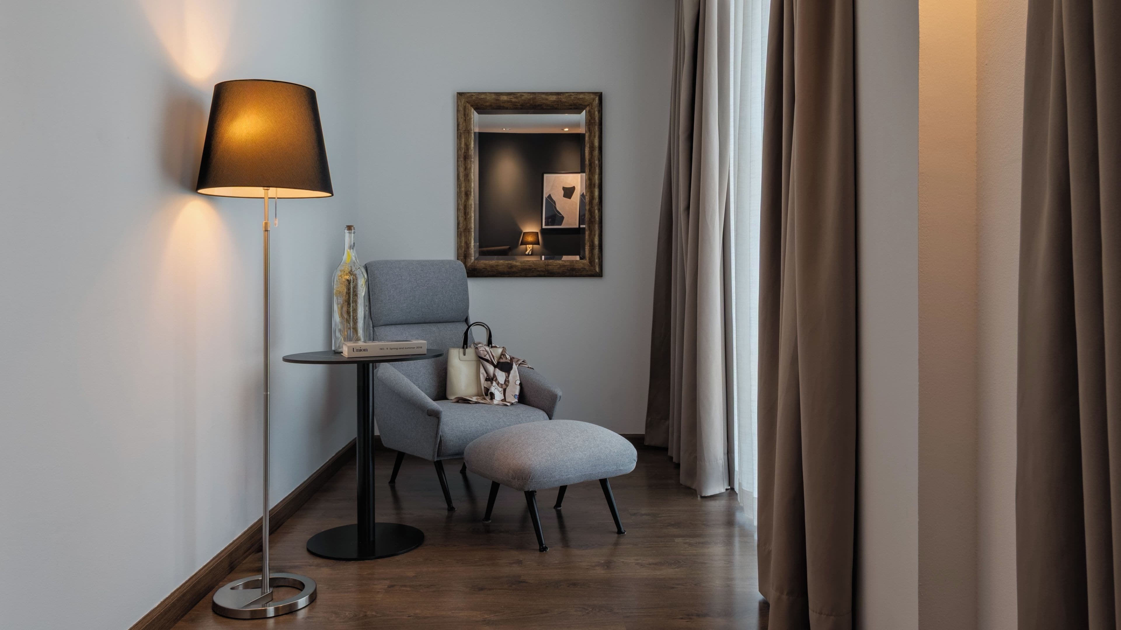 Une chambre d’hôtel moderne avec un fauteuil gris, une petite table ronde, une lampe sur pied, un miroir et une fenêtre avec des rideaux.