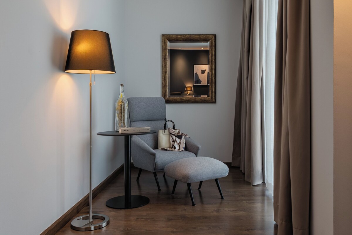 Quarto de hotel moderno com poltrona cinza, uma mesinha redonda pequena, abajur, espelho e janela com cortinas.