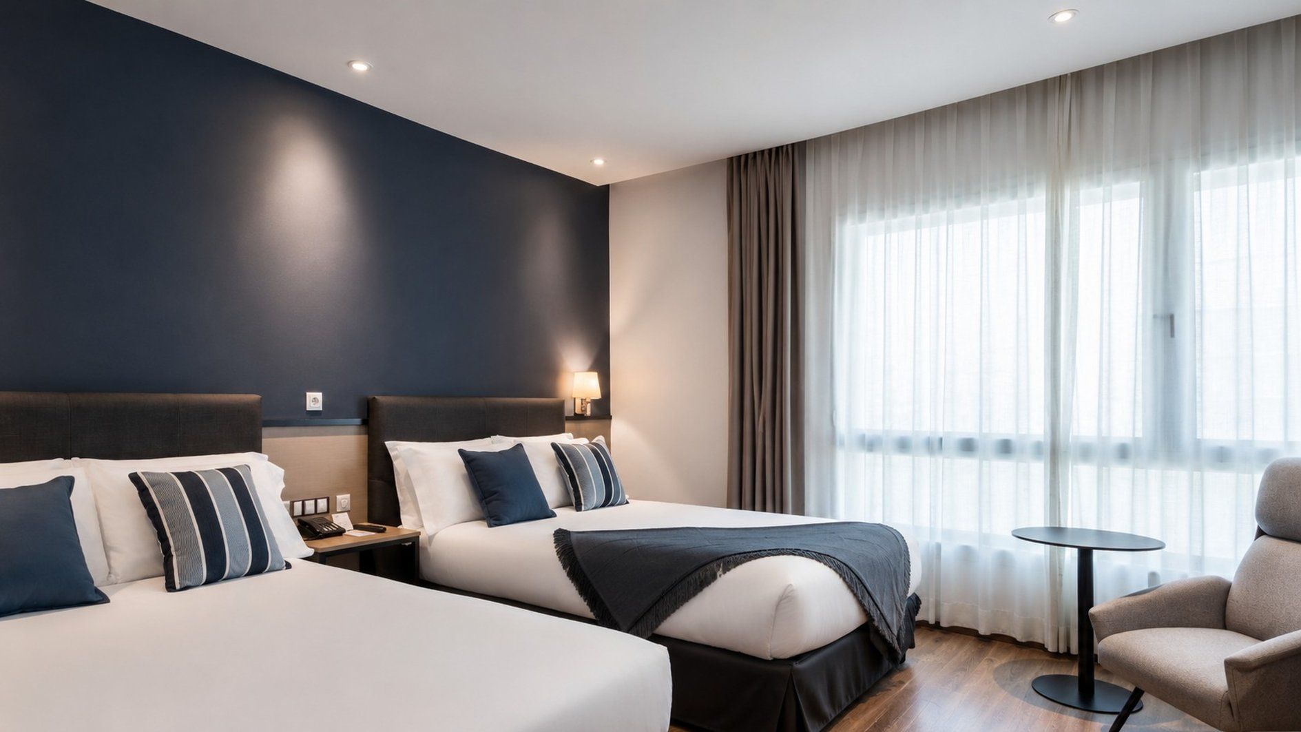 Habitación de hotel con dos camas, un escritorio, una silla y un ventanal con cortinas.