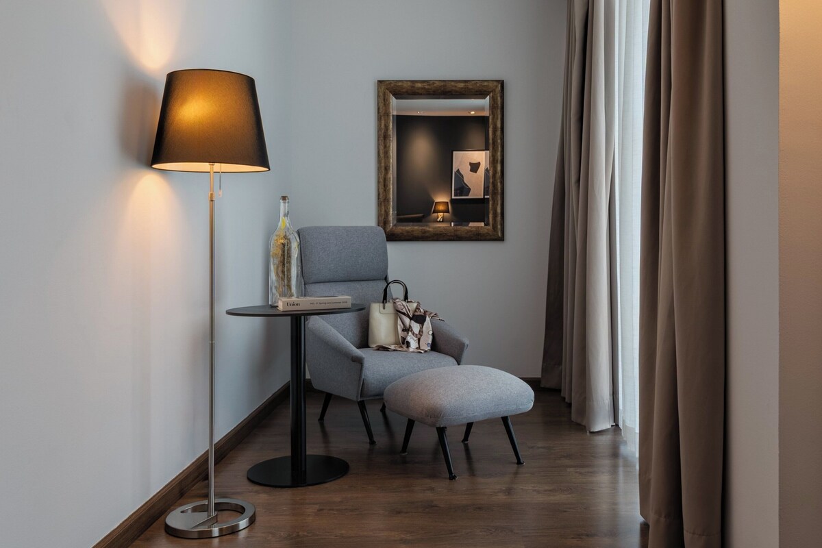 Una habitación de hotel moderna con un sillón gris, una mesita redonda pequeña, una lámpara de pie, un espejo y una ventana con cortinas.