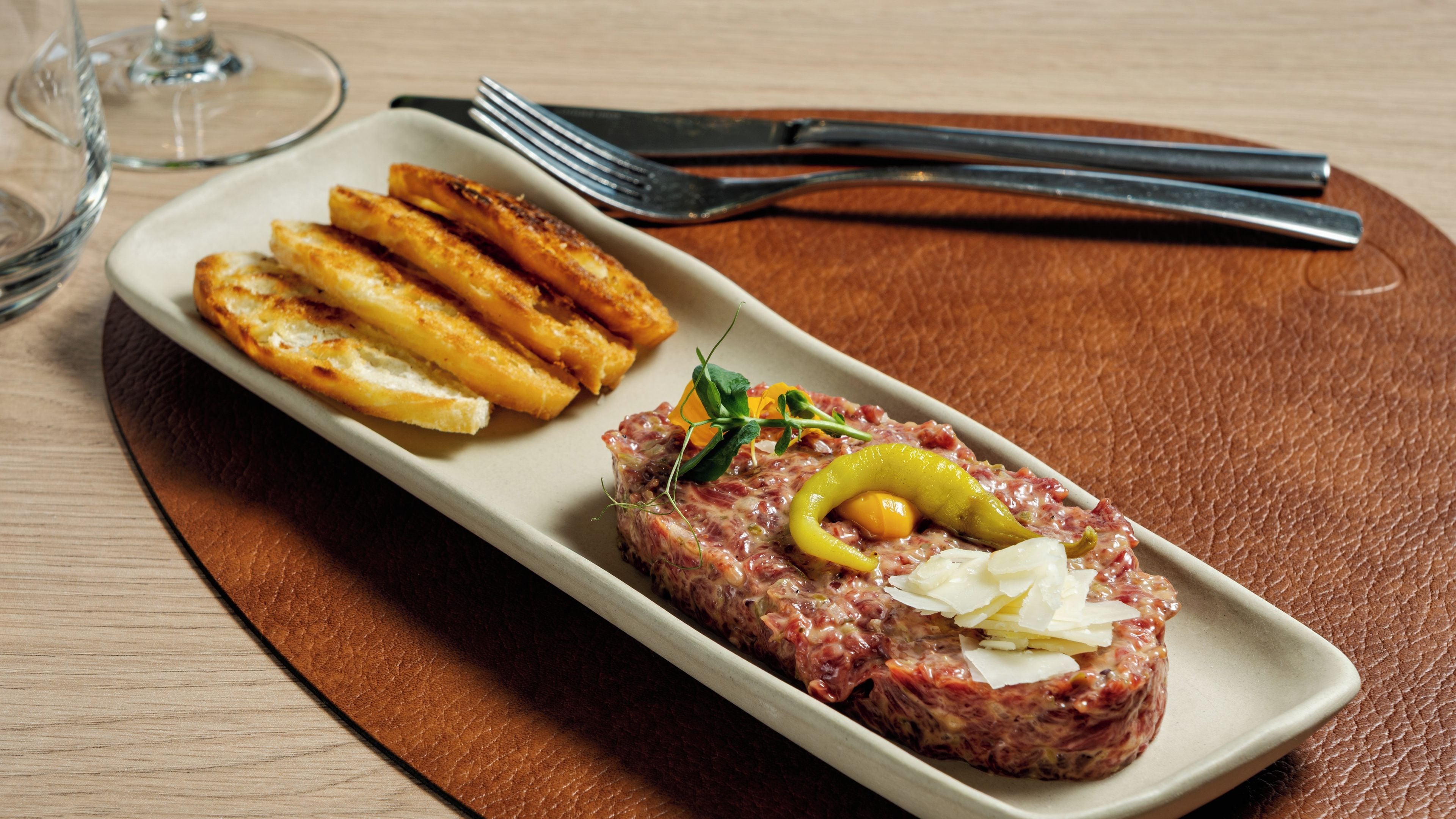 A rectangular plate with a meat dish garnished with a yellow pepper and cheese, accompanied by grilled breadsticks.