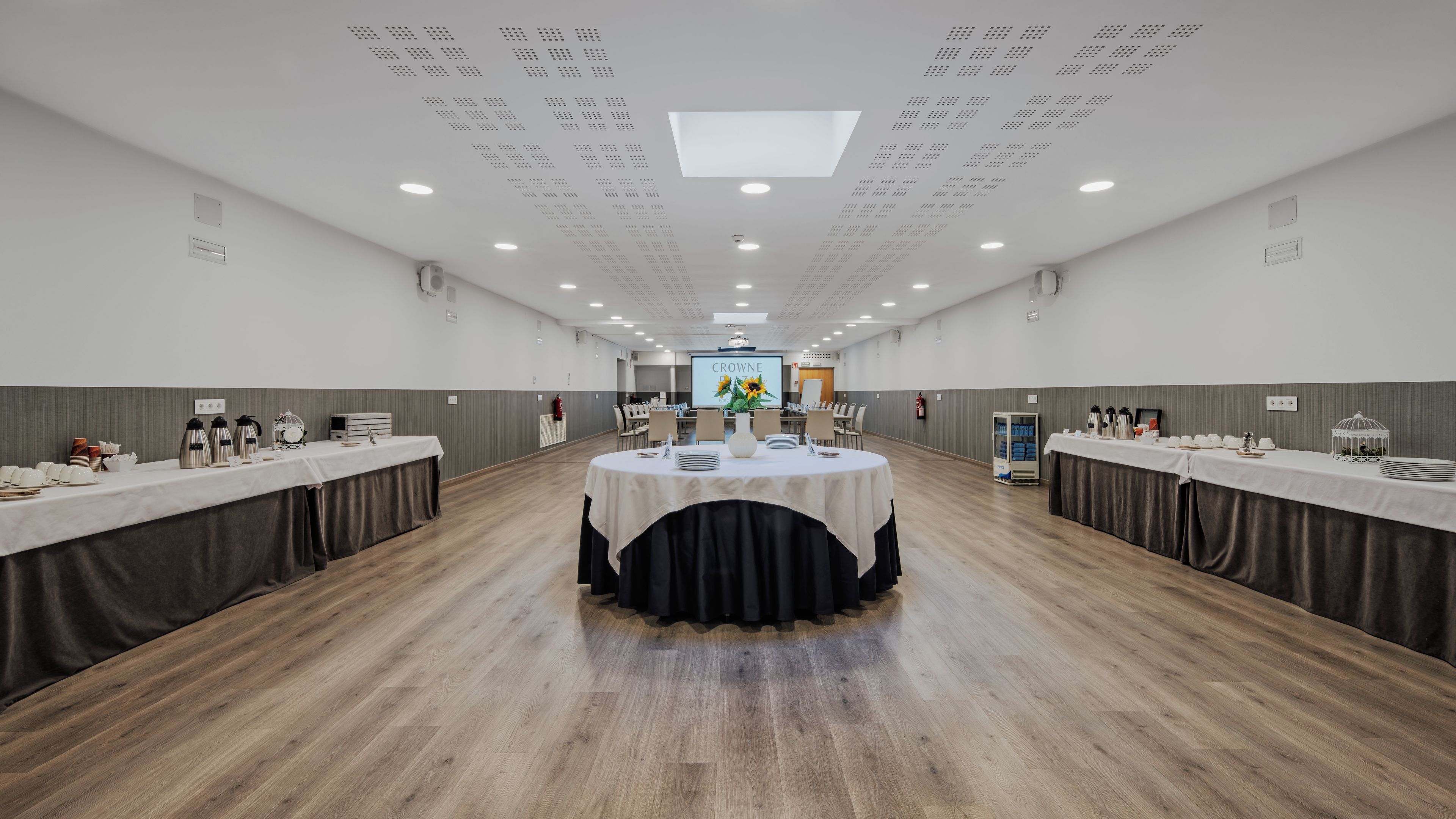 Un salón de conferencias con una mesa central, varias mesas a ambos lados y una pantalla de proyector al frente.