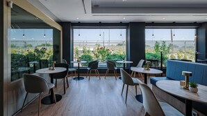 Una cafetería moderna con mesas y sillas redondas, amplios ventanales con vistas a zonas verdes y una zona de estar azul.