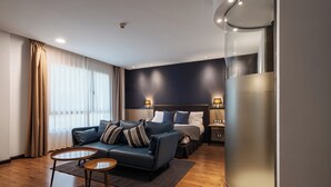 Una habitación de hotel moderna con un cabecero azul oscuro, un sofá con almohadas a rayas, una mesa de centro redonda de madera y un objeto cilíndrico grande a la derecha.