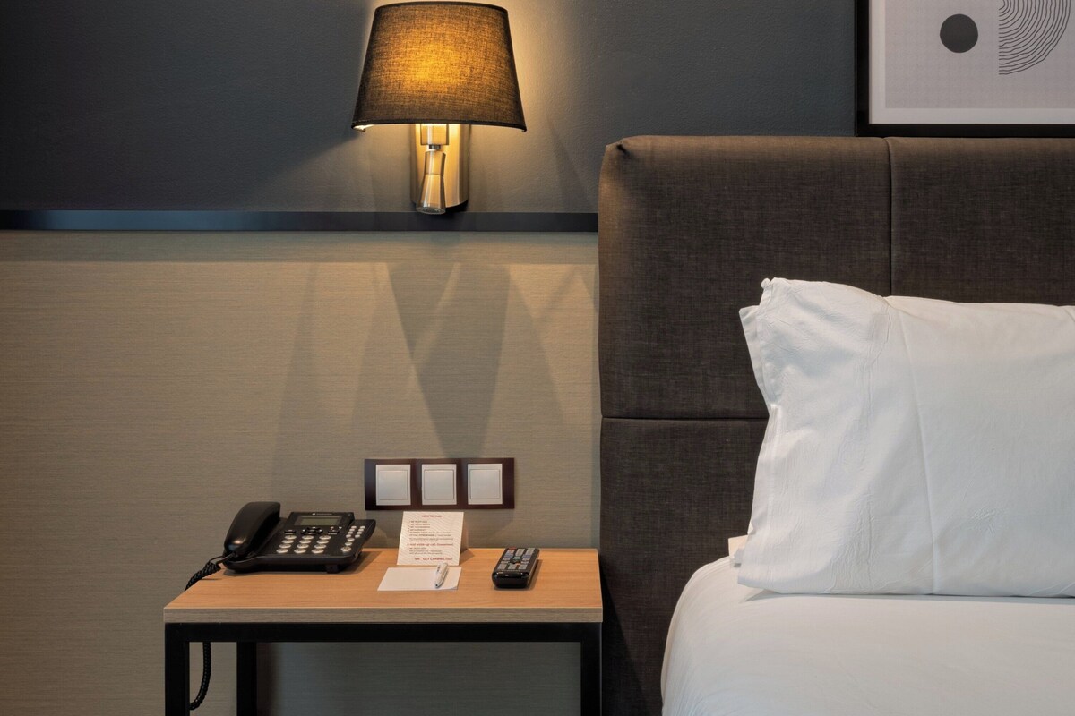 A hotel room with a bed, a nightstand with a lamp, a phone, a remote control, and a framed picture on the wall.