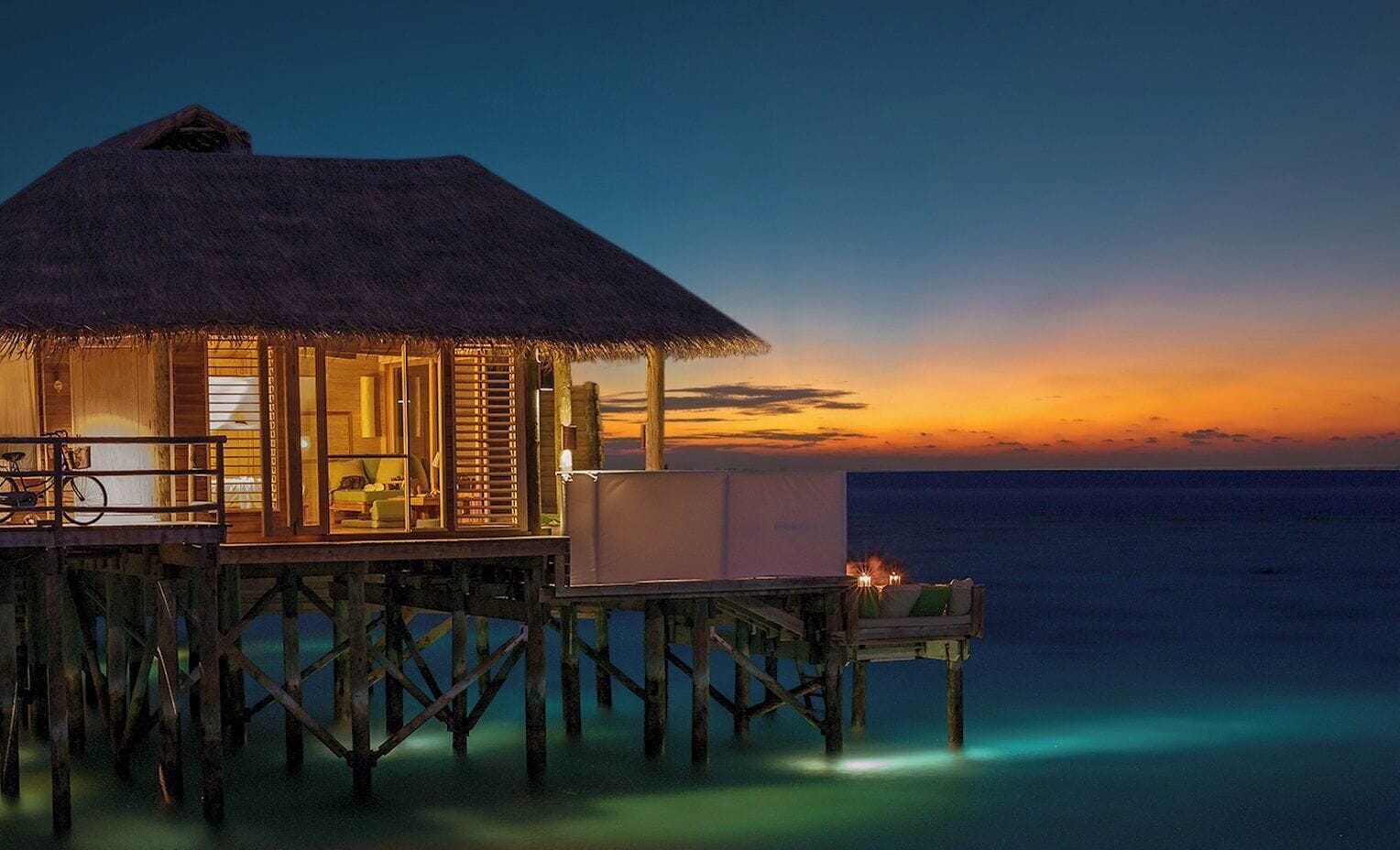 Una cabaña sobre el agua con techo de paja, un balcón, una terraza de madera y vista al océano al atardecer.