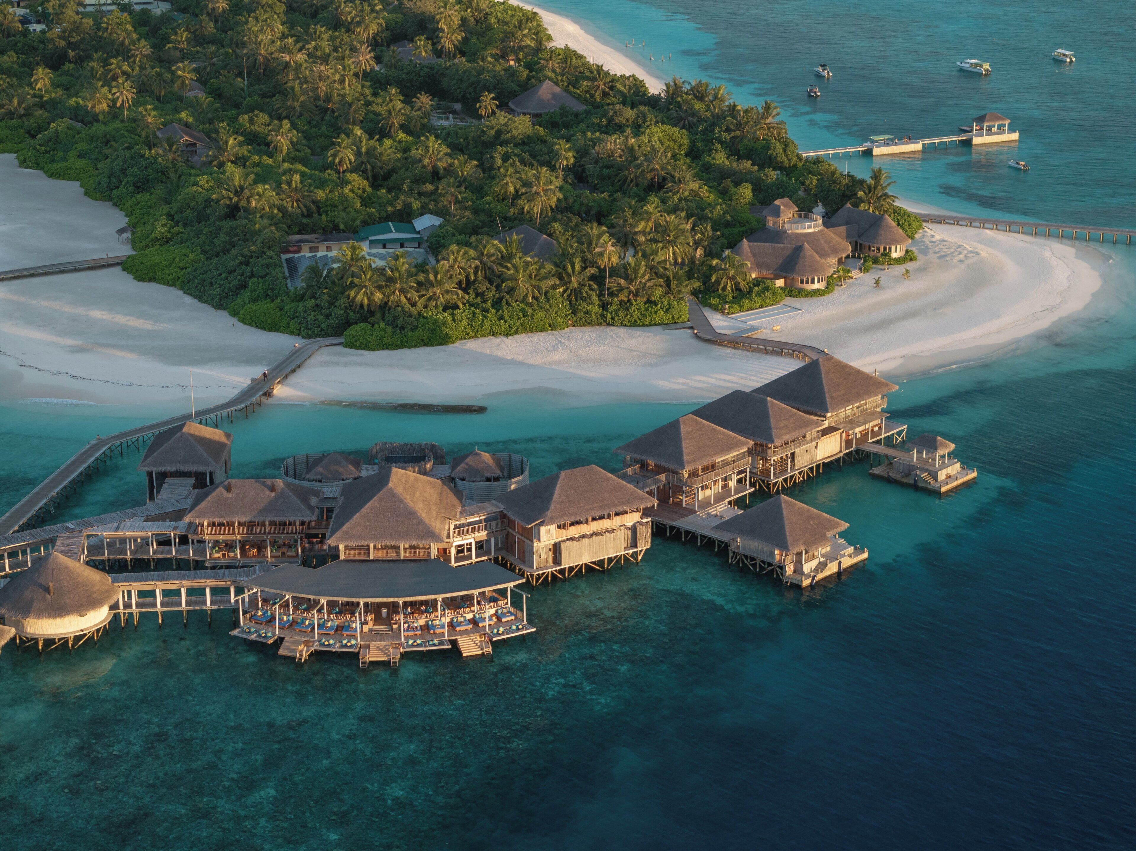 Un resort sobre el agua con varias edificaciones tipo bungalow conectadas por senderos, rodeado de aguas turquesas cristalinas y una playa de arena.