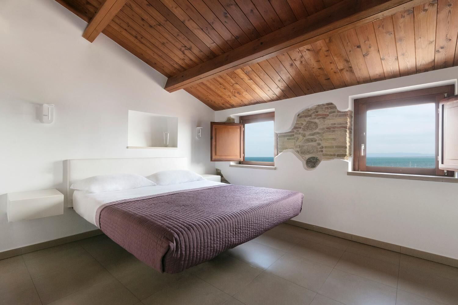 Un dormitorio con techo de madera, una cama con manta morada, pared de piedra y vista al mar por una ventana.