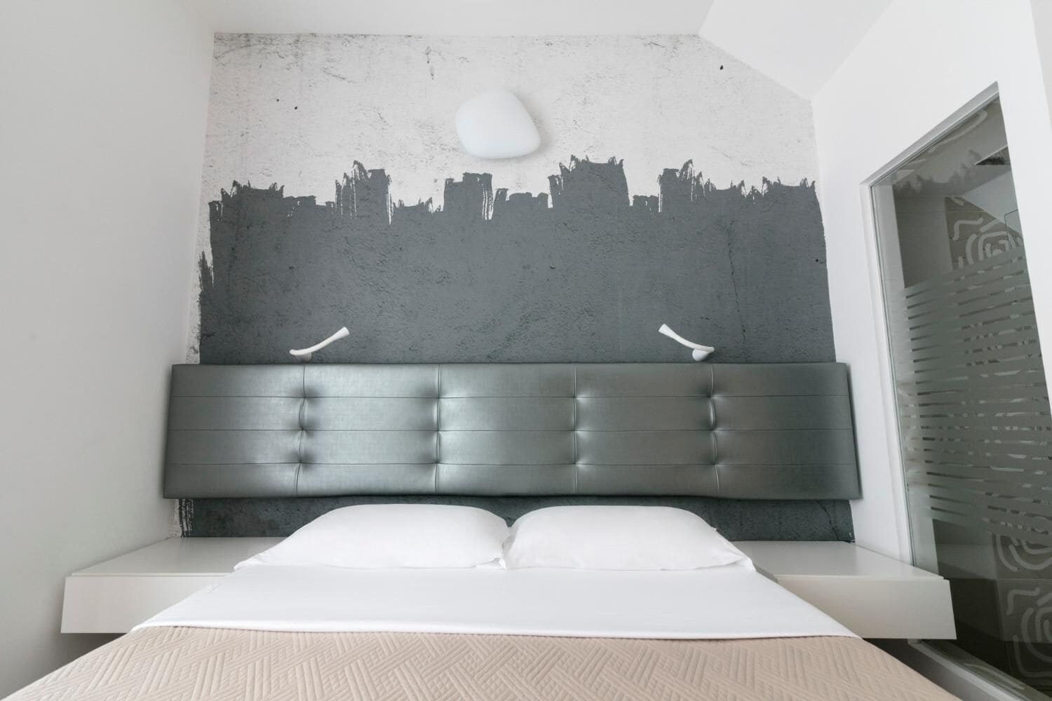 A modern bedroom with a cityscape mural on the wall, a tufted headboard, and a bed with white linens.