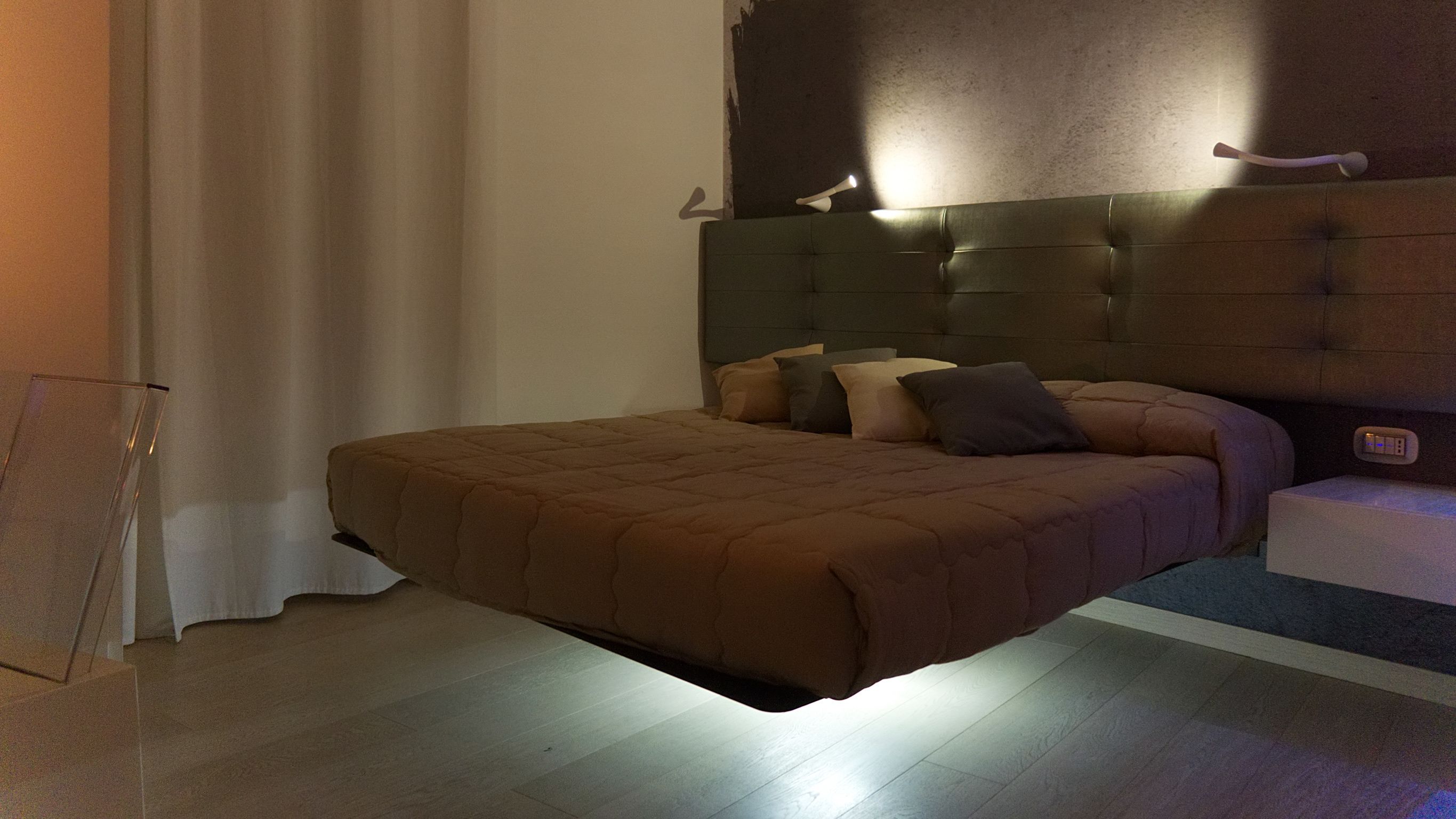 A modern bedroom with a large bed, a headboard with integrated lighting, and a bedside table with a power outlet.