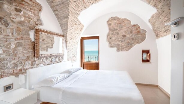 A bedroom with a stone wall, a bed, a nightstand, and a view of the sea through an open door. - Locanda Alfieri (Termoli)