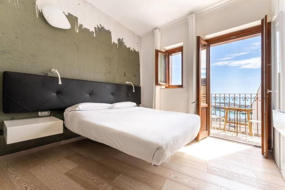 A bedroom with a bed, a bedside table, a window, and a balcony with a view of the sea.