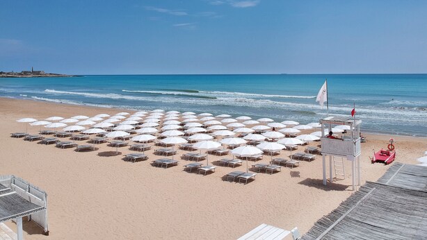 A beach with rows of white umbrellas and sun loungers, a lifeguard tower, and a clear view of the ocean. - VOI Marsa Siclà Essentia (Scicli)