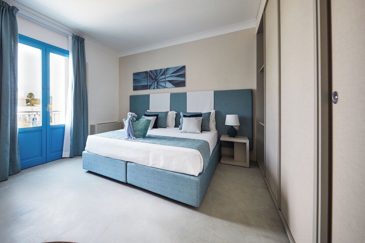 A modern hotel room with a large bed, a nightstand, a lamp, and a view of the outside through a blue-framed window.