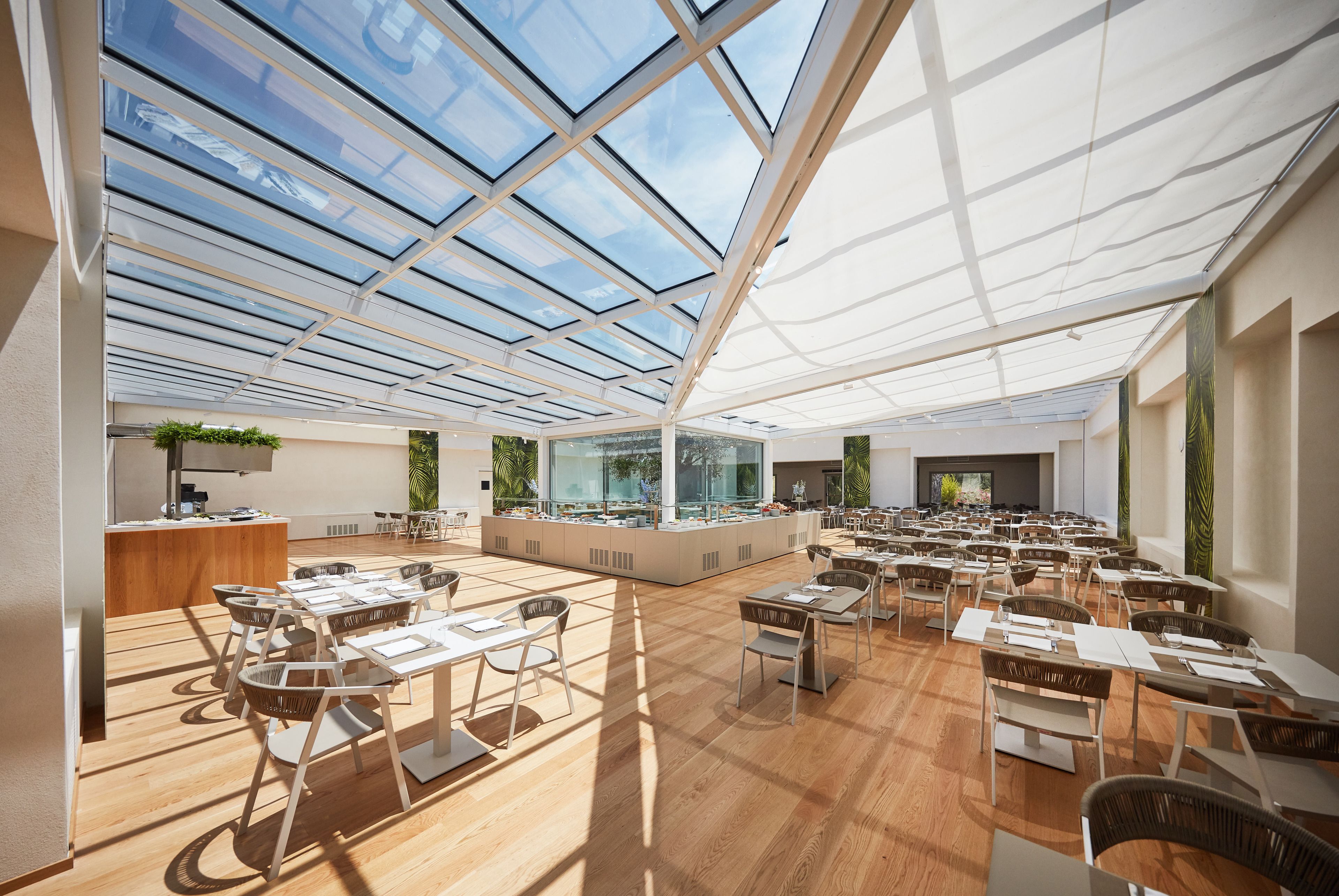 A spacious indoor area with a high ceiling and large windows, featuring tables and chairs arranged for dining or meetings.