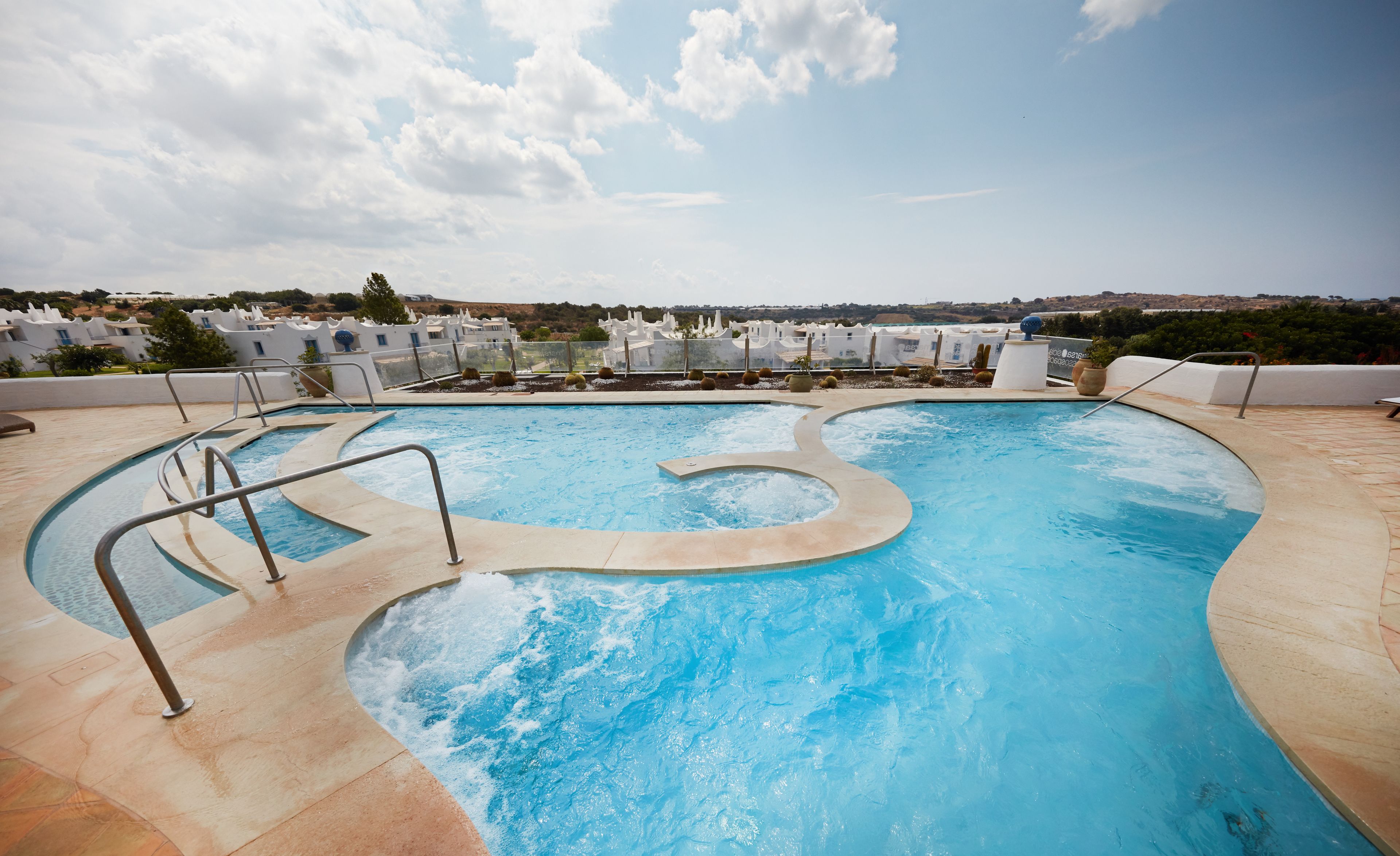 Une piscine au design original, entourée d’une terrasse carrelée et de rampes en métal. Au fond, on aperçoit des bâtiments blancs au style architectural uniforme, qui font probablement partie d’un complexe hôtelier ou d’un hôtel.
