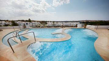 Une piscine au design original, entourée d’une terrasse carrelée et de rampes en métal. Au fond, on aperçoit des bâtiments blancs au style architectural uniforme, qui font probablement partie d’un complexe hôtelier ou d’un hôtel.