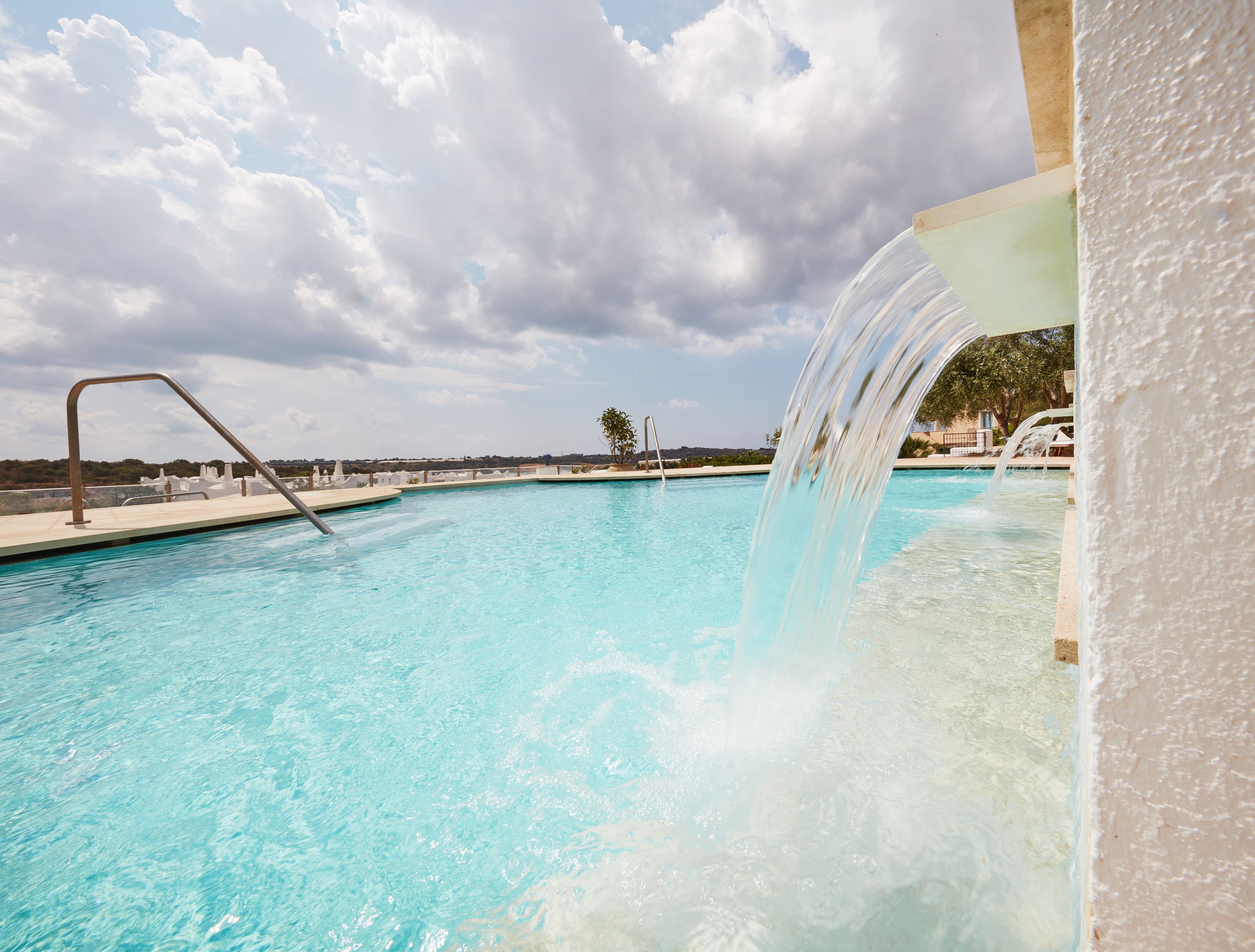 A swimming pool with a water feature, clear blue water, and a cloudy sky.