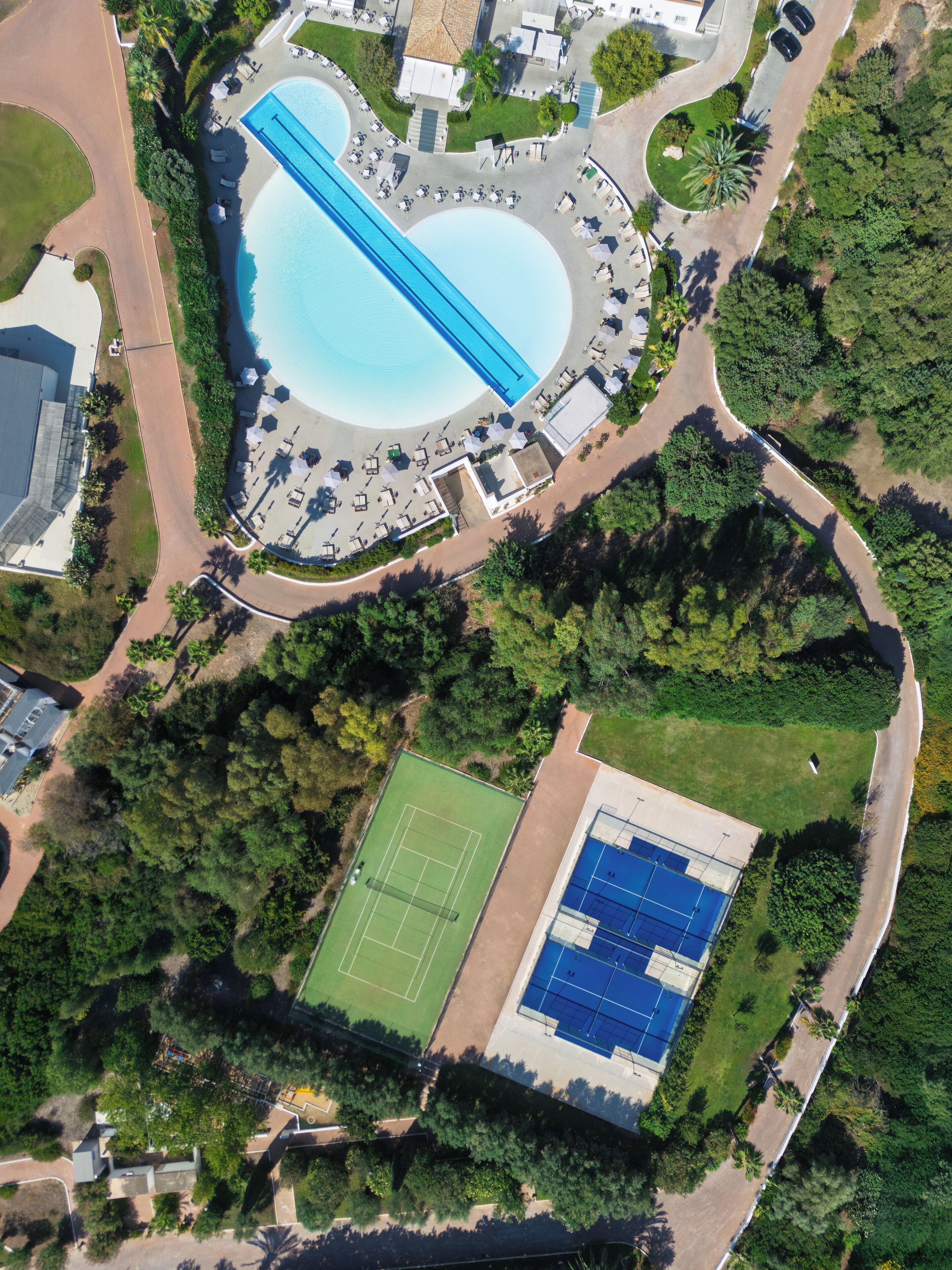 An aerial view of a resort with a large swimming pool, tennis courts, and a landscaped area with pathways and seating.