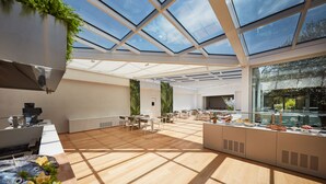 A spacious room with a high ceiling and skylights, featuring a long counter with food and drinks, wooden flooring, and a view of greenery outside. - VOI Marsa Siclà Essentia (Scicli)