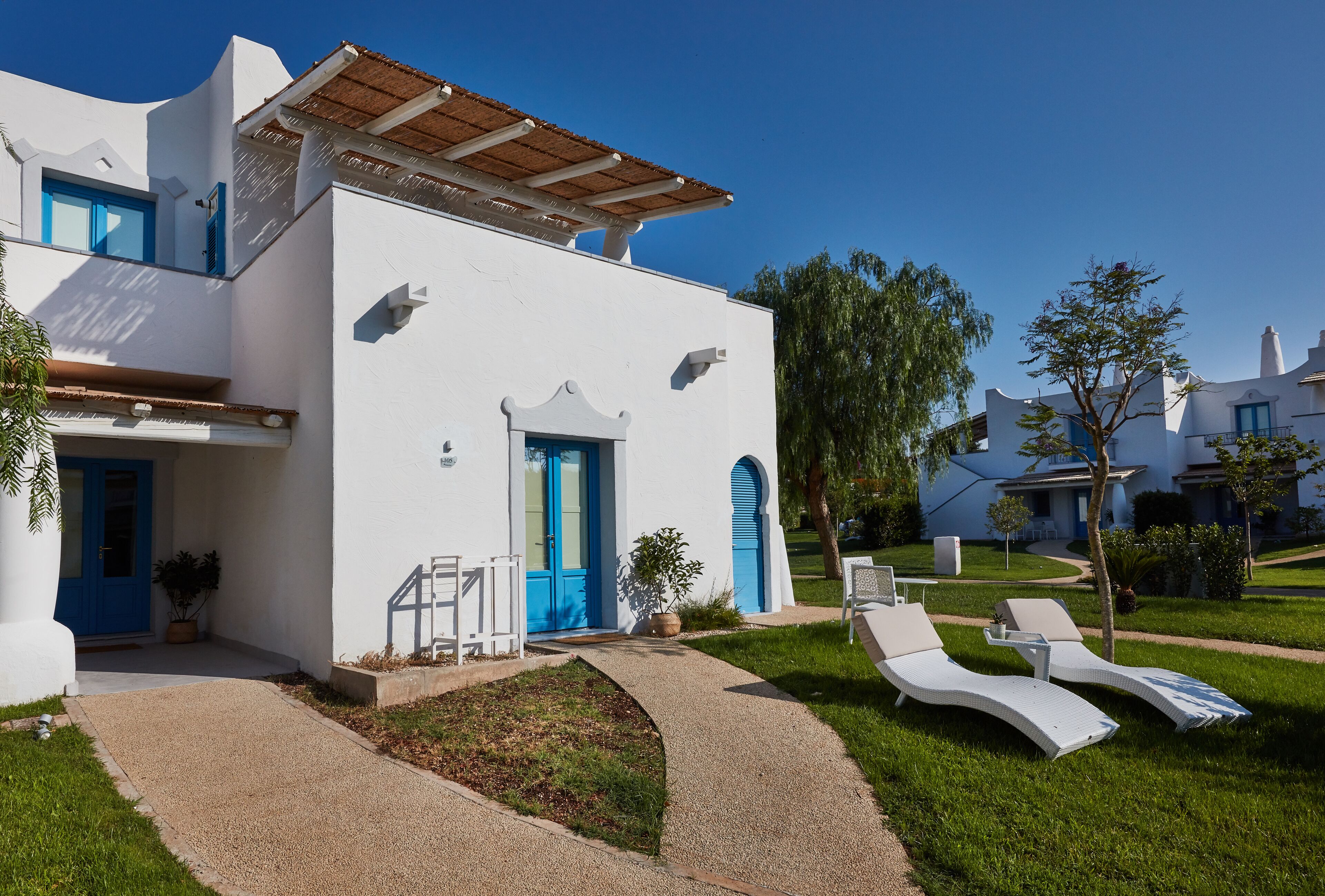 A white building with a blue door and windows, a small garden with a pathway, and two white lounge chairs.
