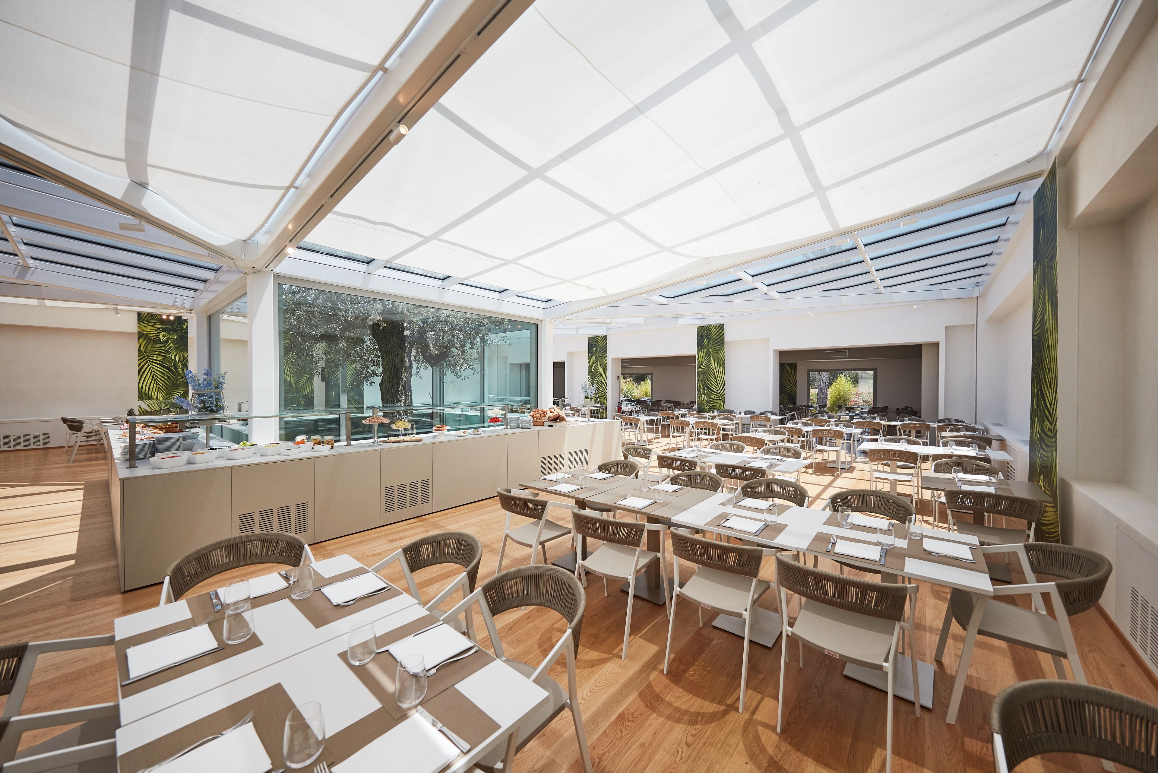 Amplia zona de comedor interior con techo alto y ventanas grandes, que presenta una isla central con una fuente de agua y asientos distribuidos a su alrededor.