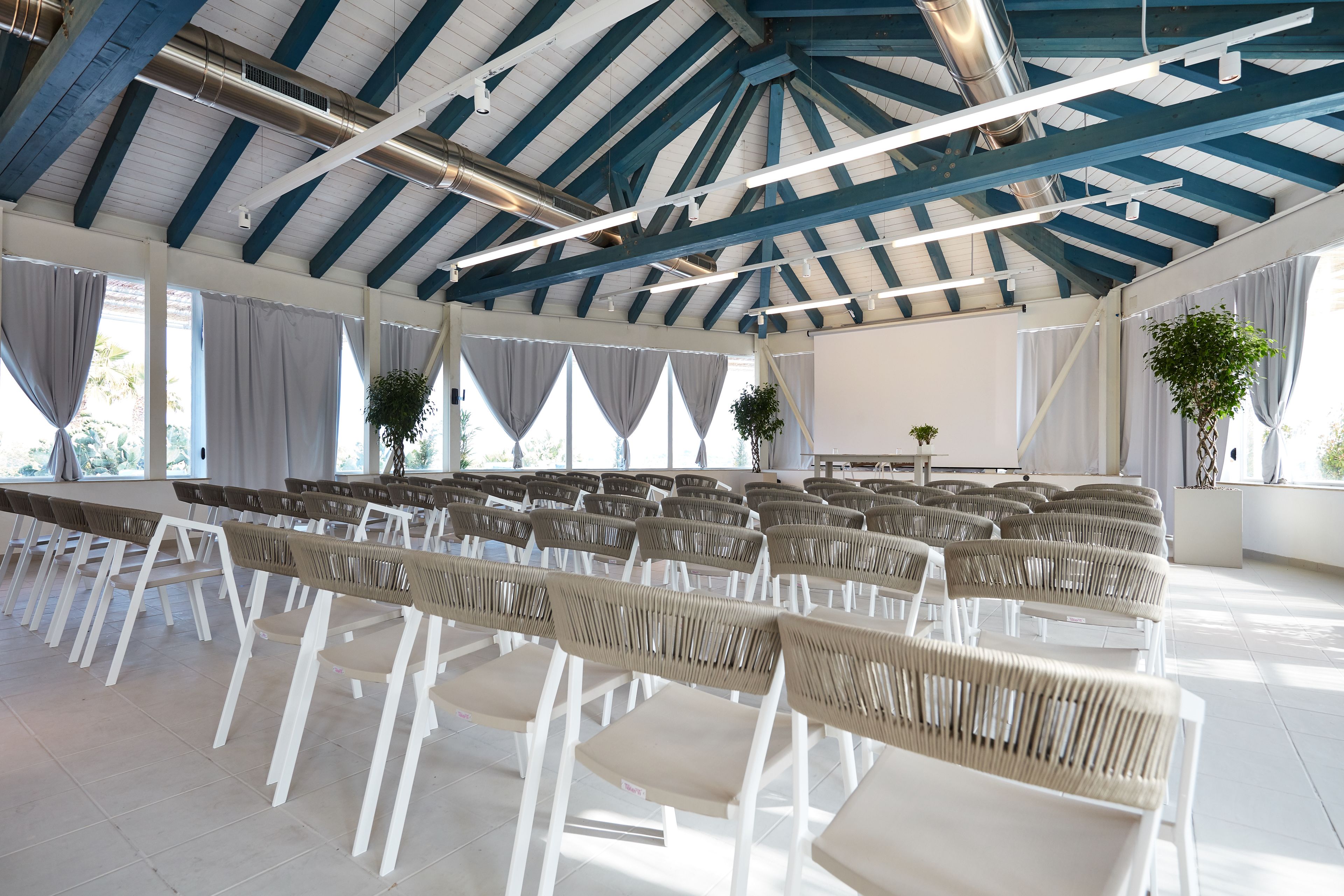 A spacious indoor venue with rows of white chairs and tables, a high ceiling with exposed beams, and large windows allowing natural light.