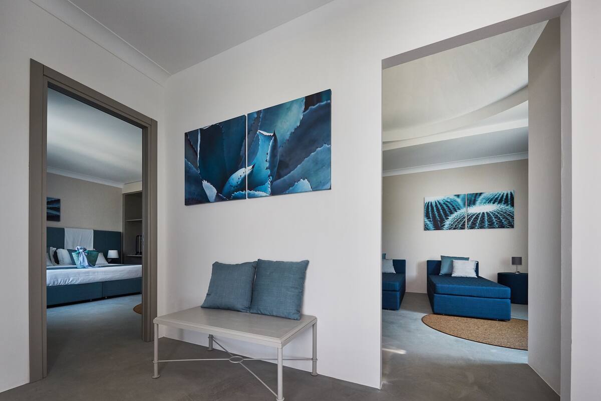 A modern hotel room with a large bed, a bench, and two blue sofas. There are abstract paintings on the walls.