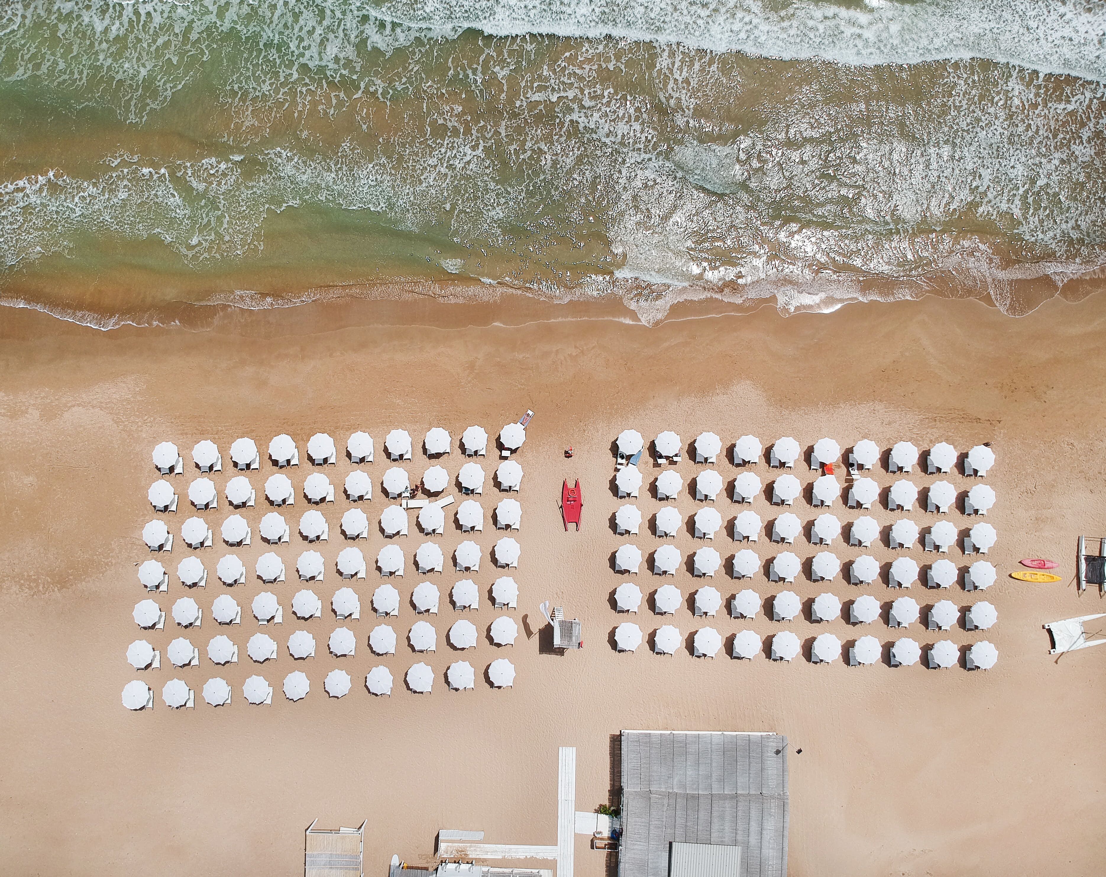 Vista aérea de una playa con hileras de sillas y sombrillas blancas, una silla de salvavidas roja y una estructura de madera.