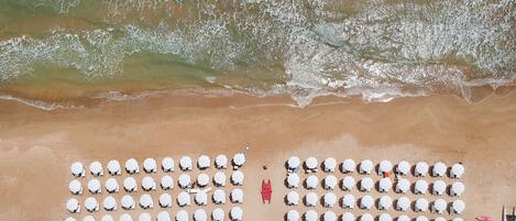 Vista aérea de una playa con hileras de sillas y sombrillas blancas, una silla de salvavidas roja y una estructura de madera.