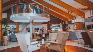 A spacious lobby with a wooden floor, a central seating area with armchairs, a glass railing, and a bar area in the background. - Draycote Hotel and Whitefields Golf Club (Rugby)