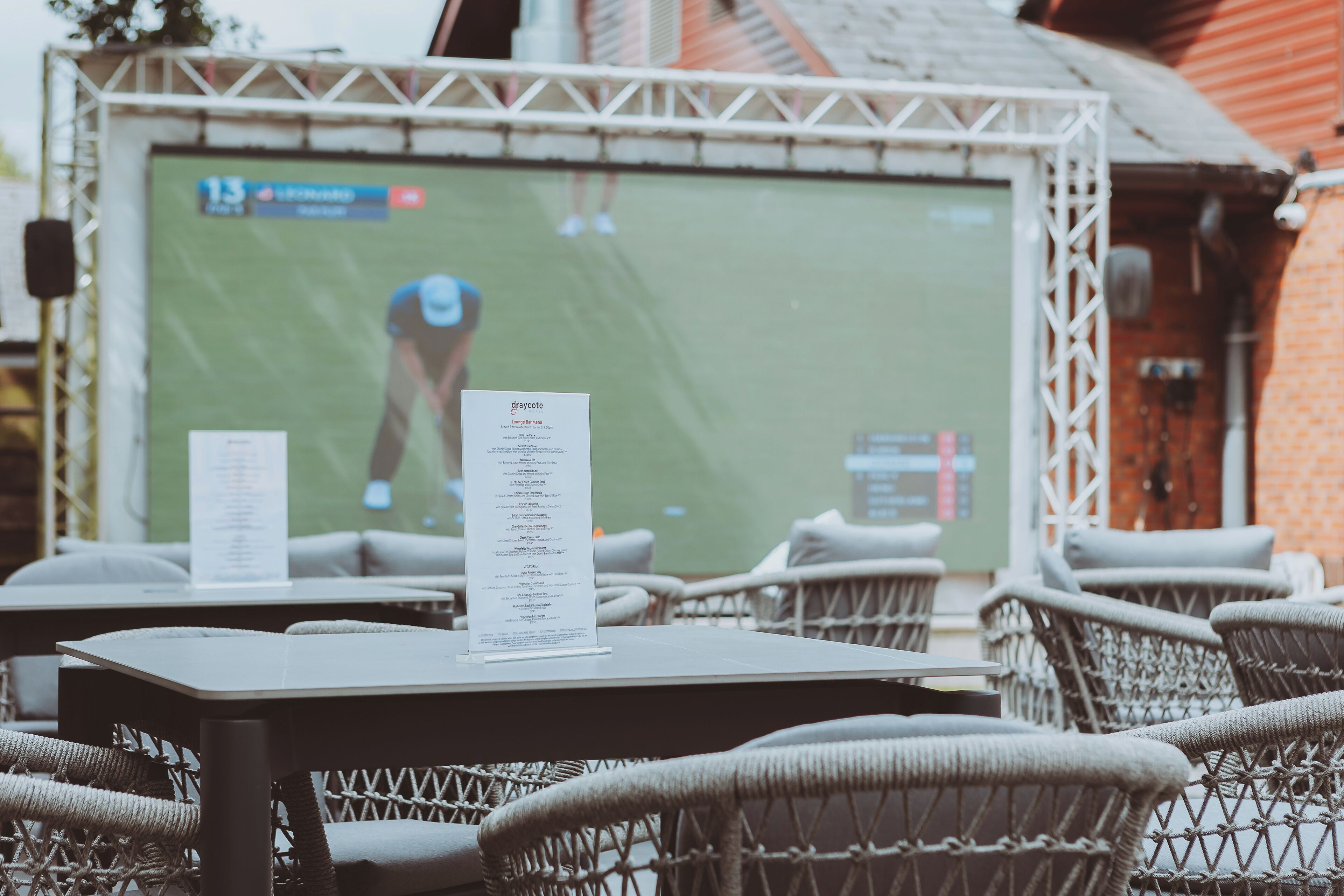 Outdoor seating with wicker chairs and tables, a menu stand, and a sports screen in the background.