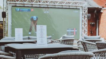 Outdoor seating with wicker chairs and tables, a menu stand, and a sports screen in the background.