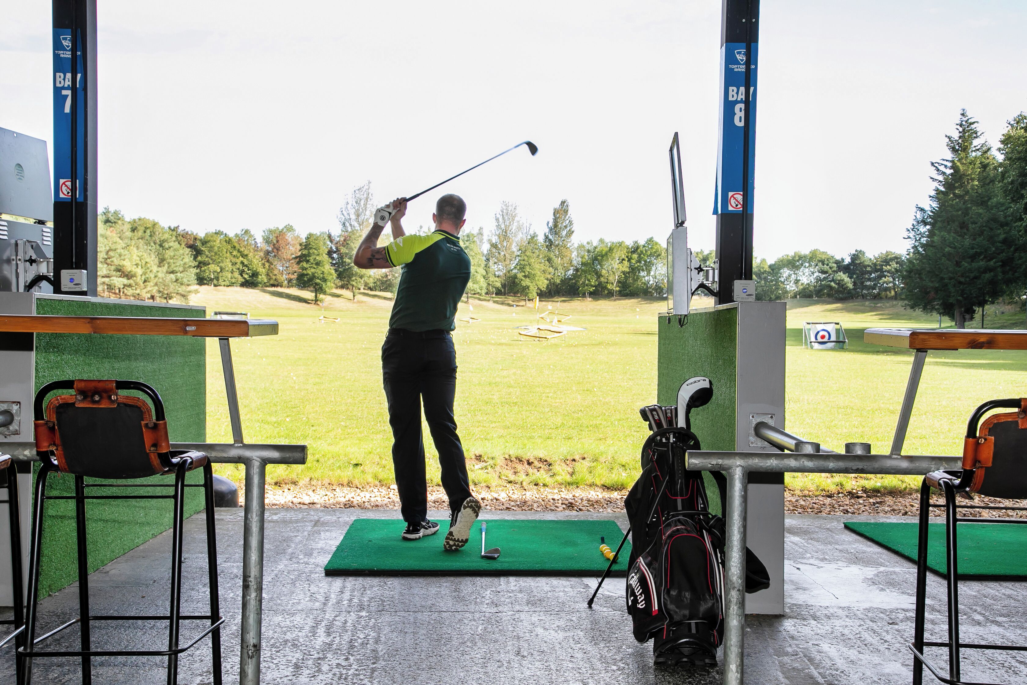 A golf driving range with a golfer practicing on a green mat, a golf bag, and a target in the background.