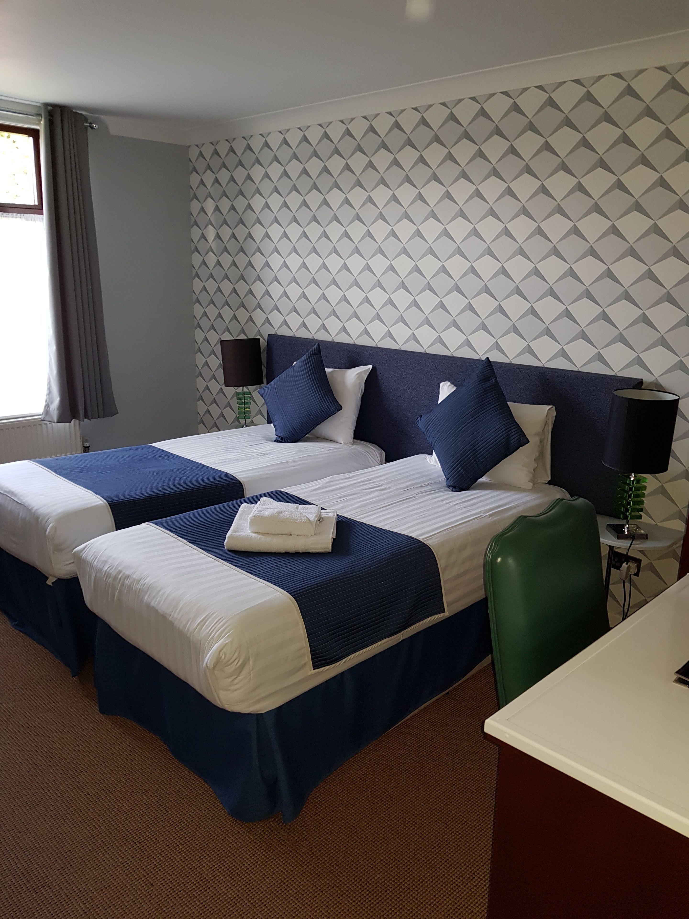 A hotel room with two beds, a green chair, a desk, and a patterned wall.