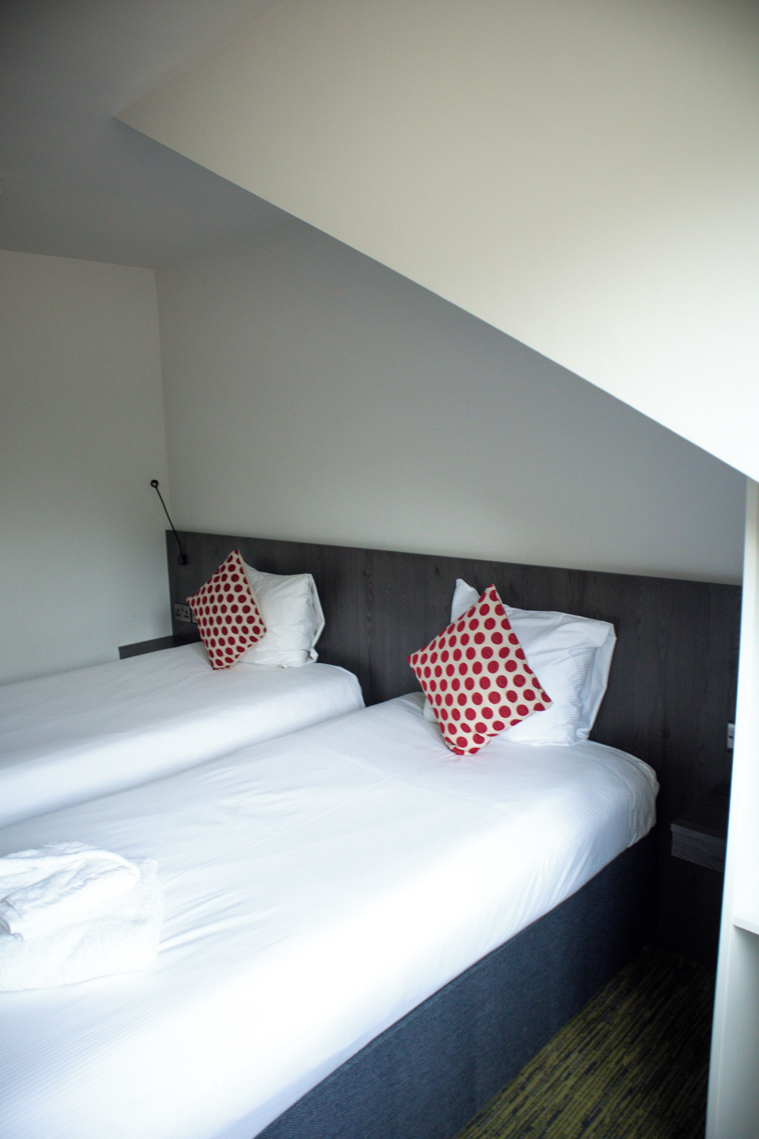 Una habitaci ón de hotel con dos camas individuales, cada una con ropa de cama blanca y almohadas rojas con lunares. La habitación tiene un cabecero de madera y una mesita de noche pequeña con una lámpara.