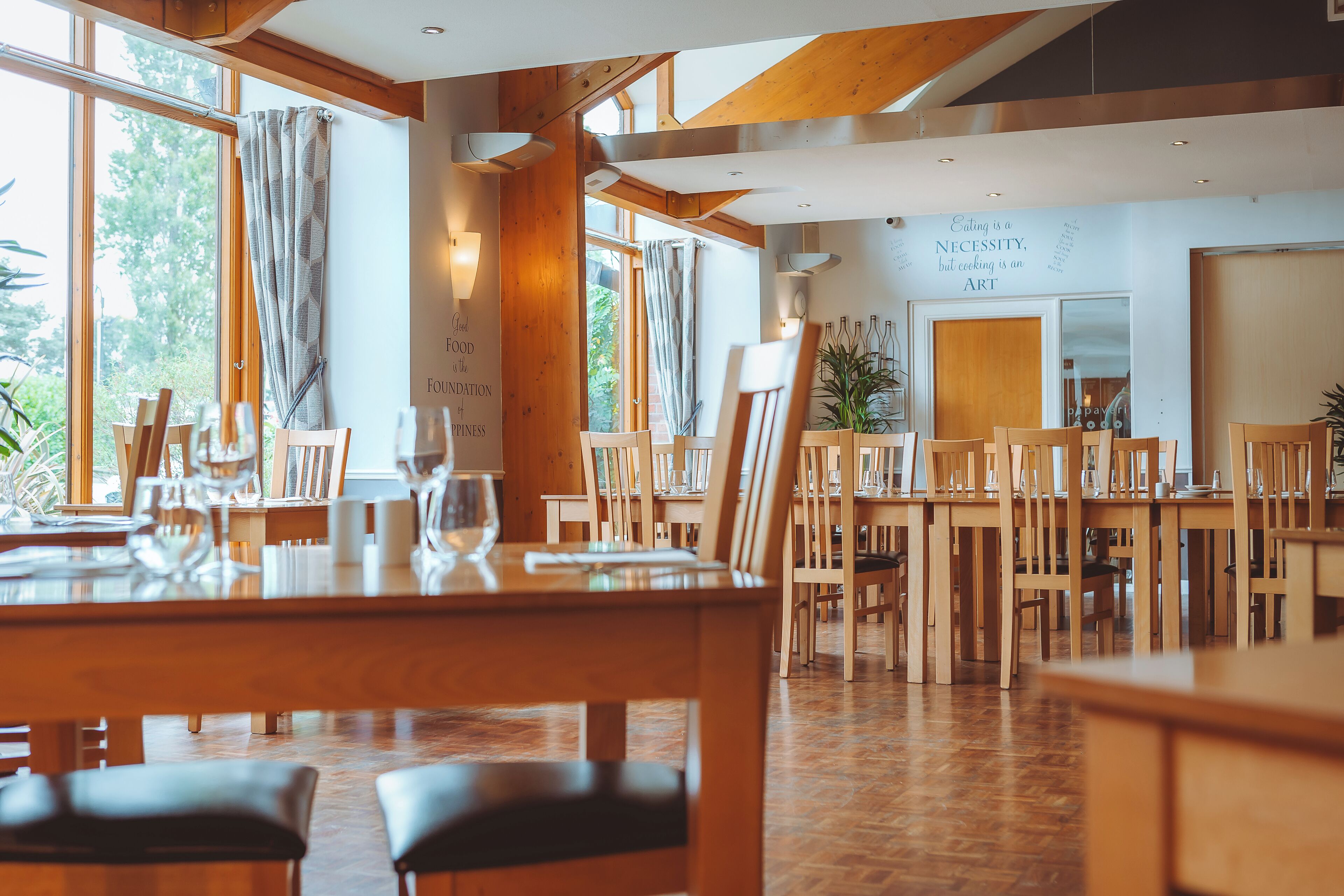 A restaurant with wooden tables and chairs, large windows, and a wall with a quote about food.