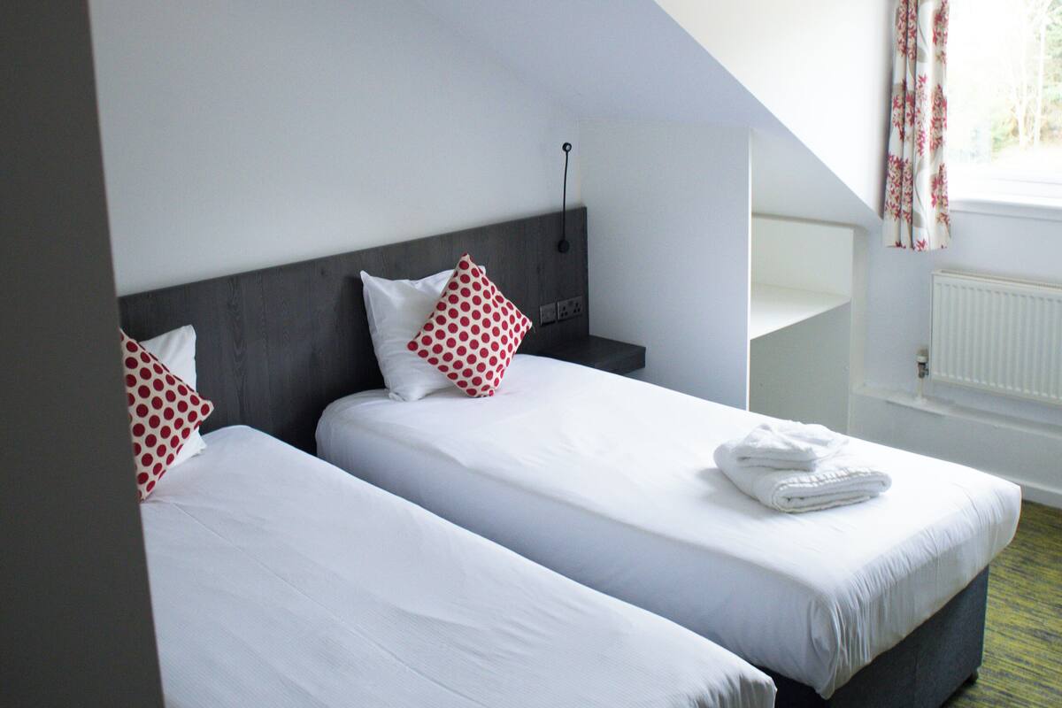 A hotel room with two beds, a ceiling light, a radiator, and a window with curtains.