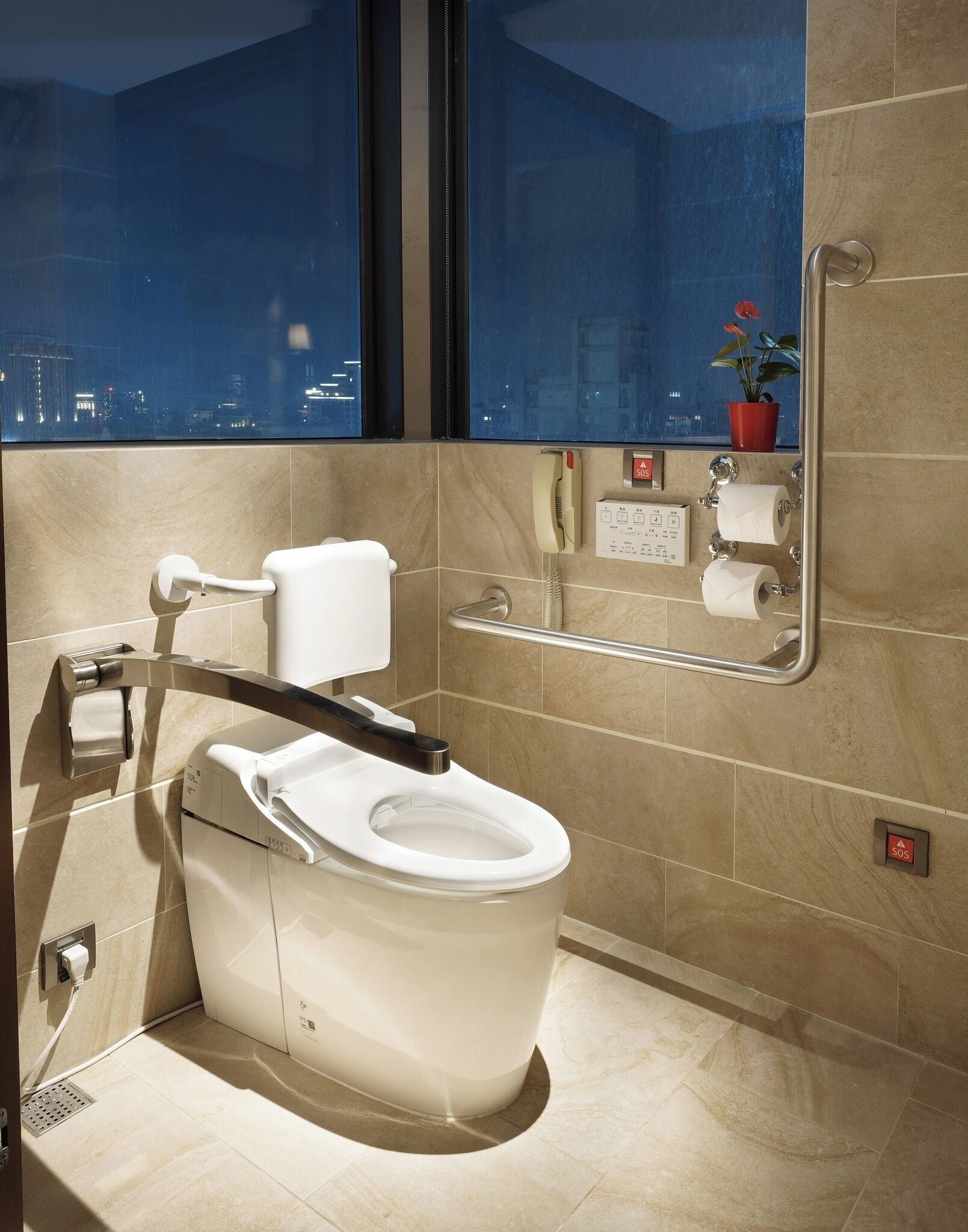 A modern bathroom with a toilet, handrails, a call button, and a window with a city view.