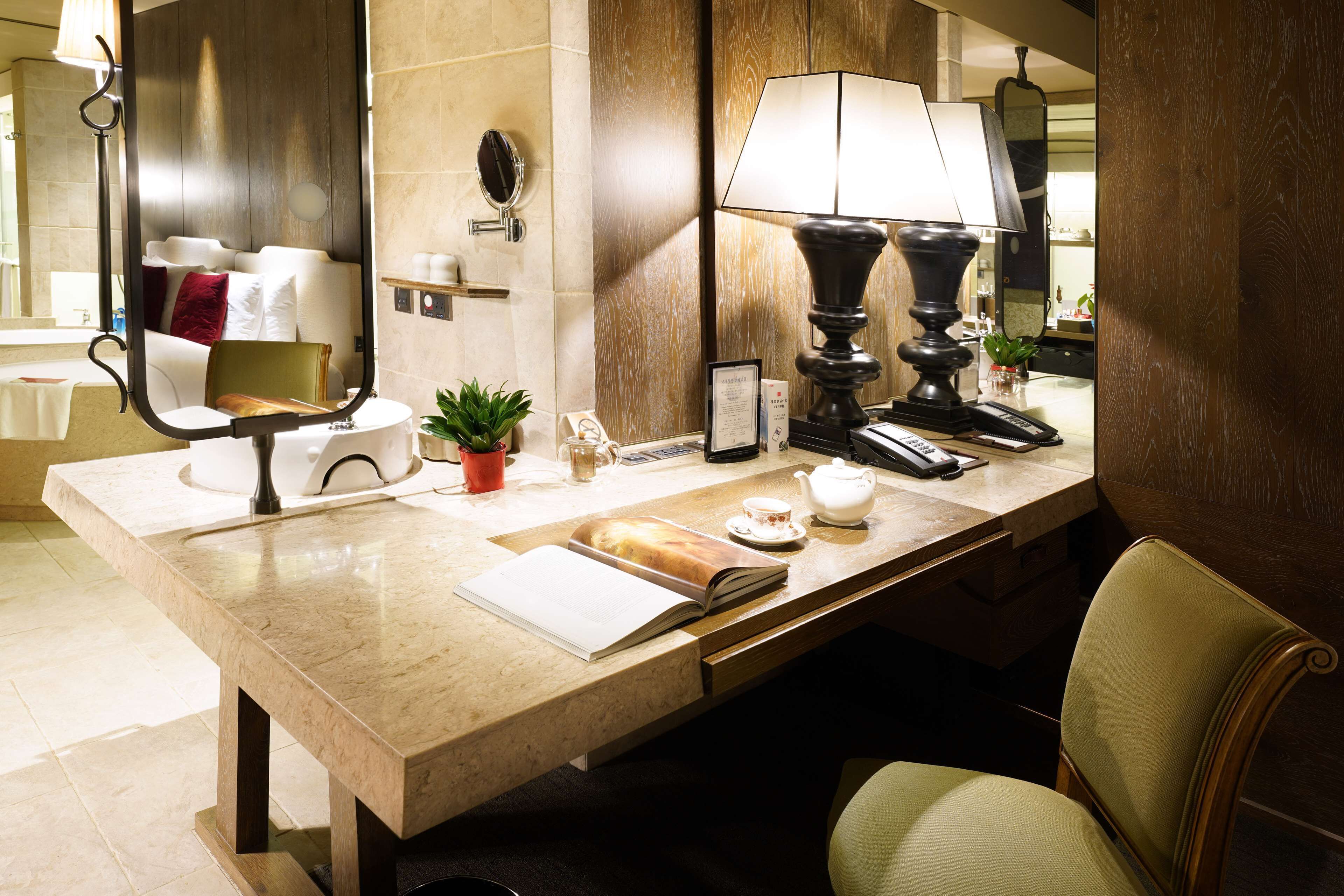 A well-lit hotel bathroom with a large mirror, a sink, a towel rack, a small plant, and a bedside table with a lamp and a book.