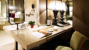 A well-lit hotel bathroom with a large mirror, a sink, a towel rack, a small plant, and a bedside table with a lamp and a book.