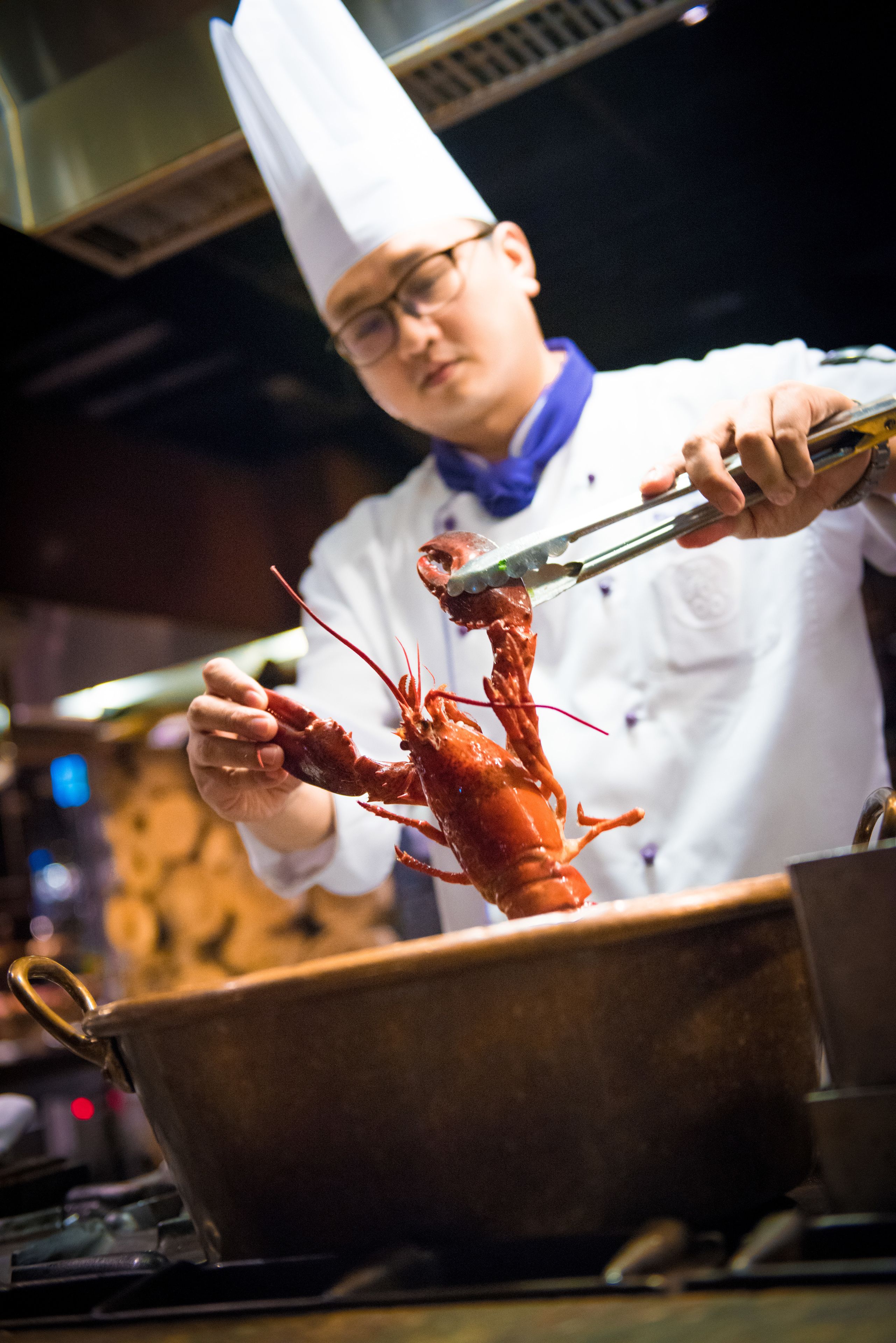 A chef holding a lobster with tongs, in a kitchen setting.