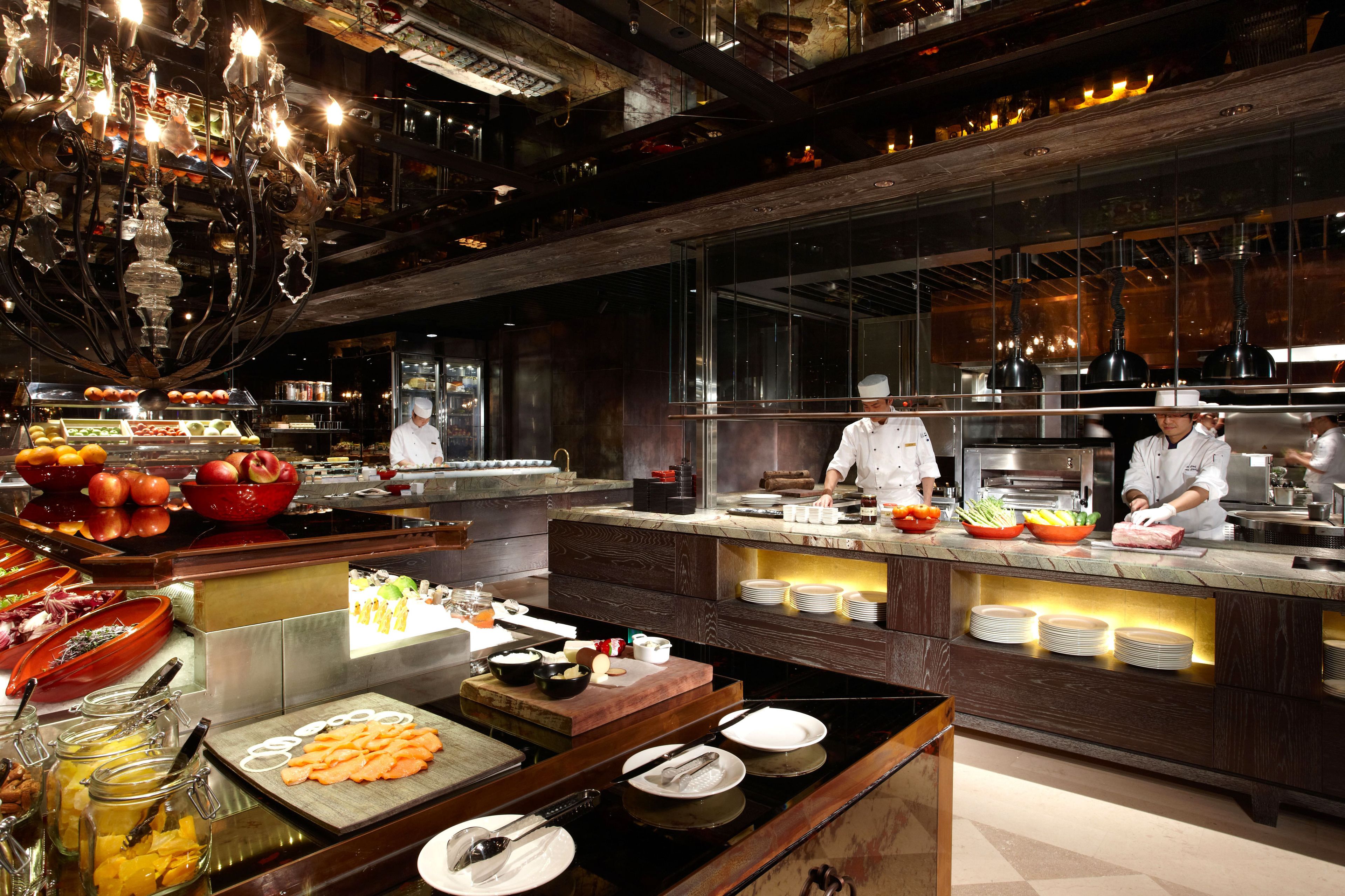 Une cuisine moderne où des chefs préparent les plats, un buffet proposant divers mets et un lustre.