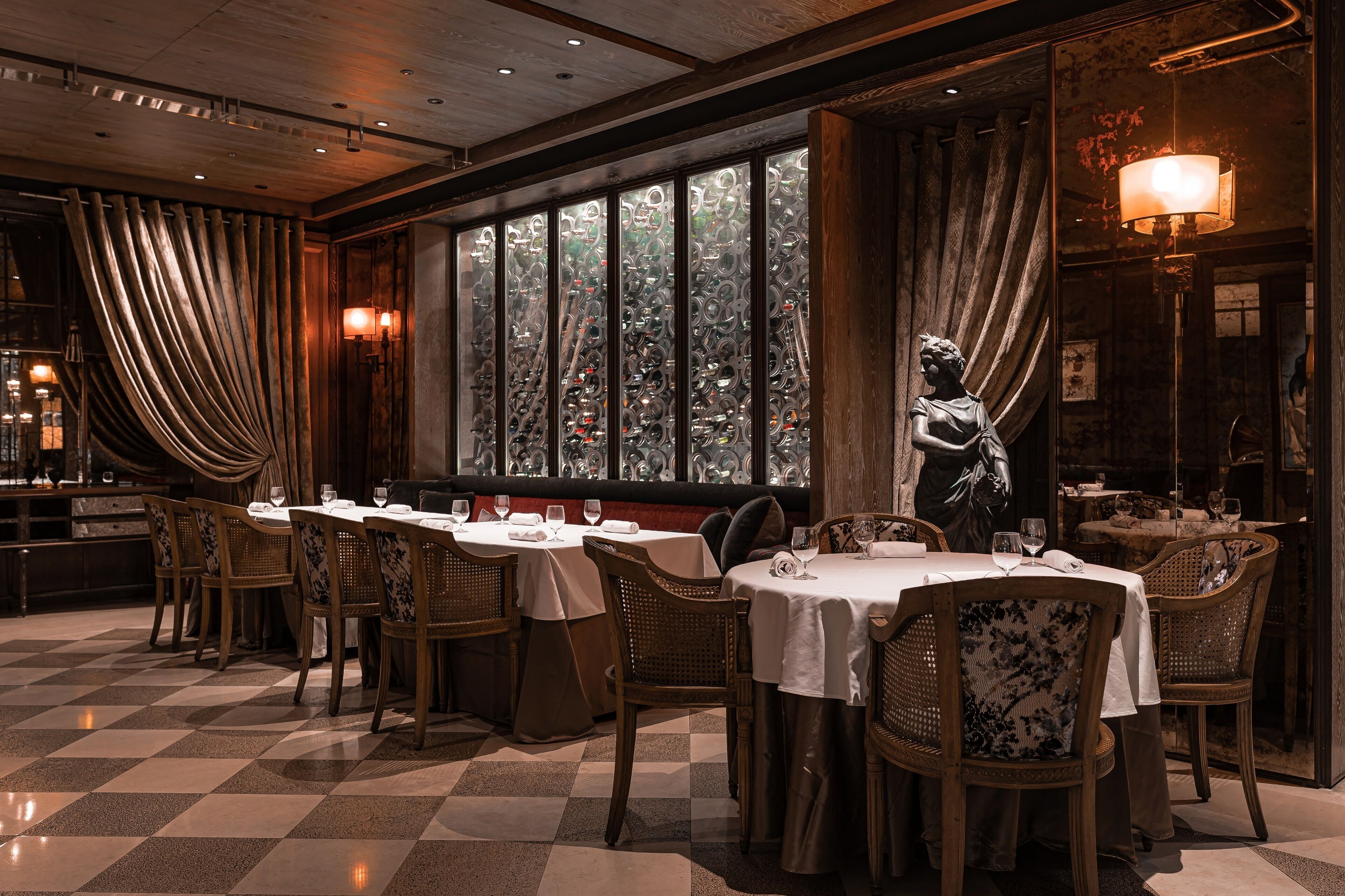 A restaurant with wooden tables and chairs, large windows with curtains, and a statue.
