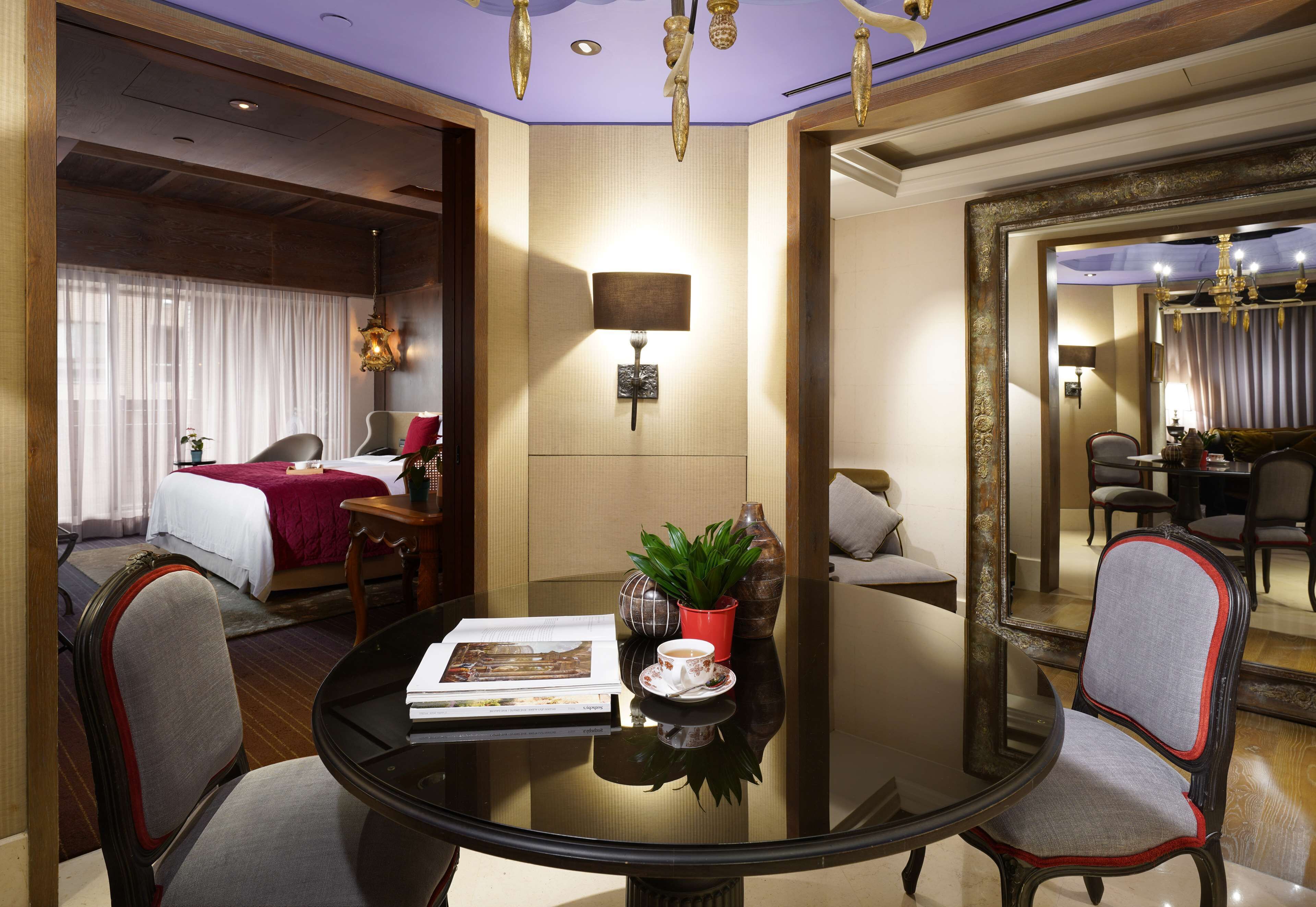 A hotel room with a round table, two chairs, a bed with a red blanket, a potted plant, and a wall-mounted lamp.