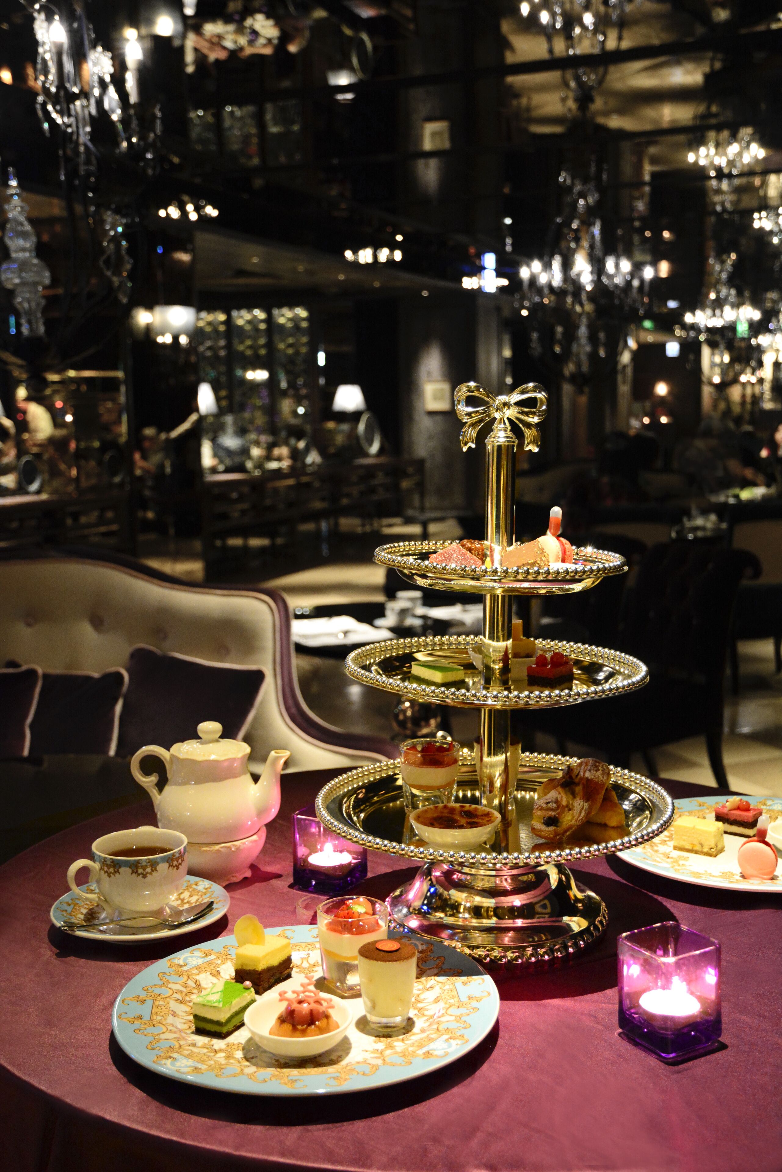 A multi-tiered dessert stand with various sweets and a teapot on a table, surrounded by candles and a sofa in a dimly lit room with chandeliers.