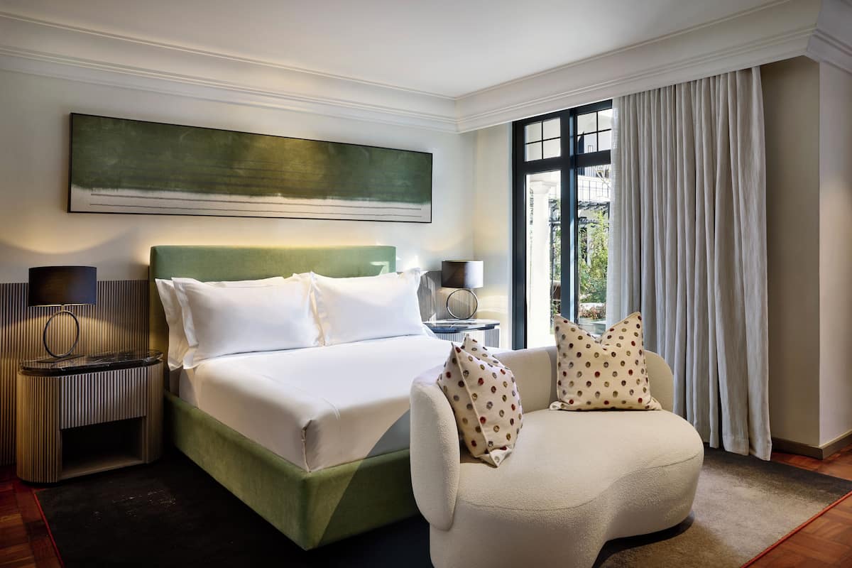 Une chambre d’hôtel avec un lit, une chaise, une table de chevet, une lampe, une grande fenêtre avec des rideaux et un tableau au mur.