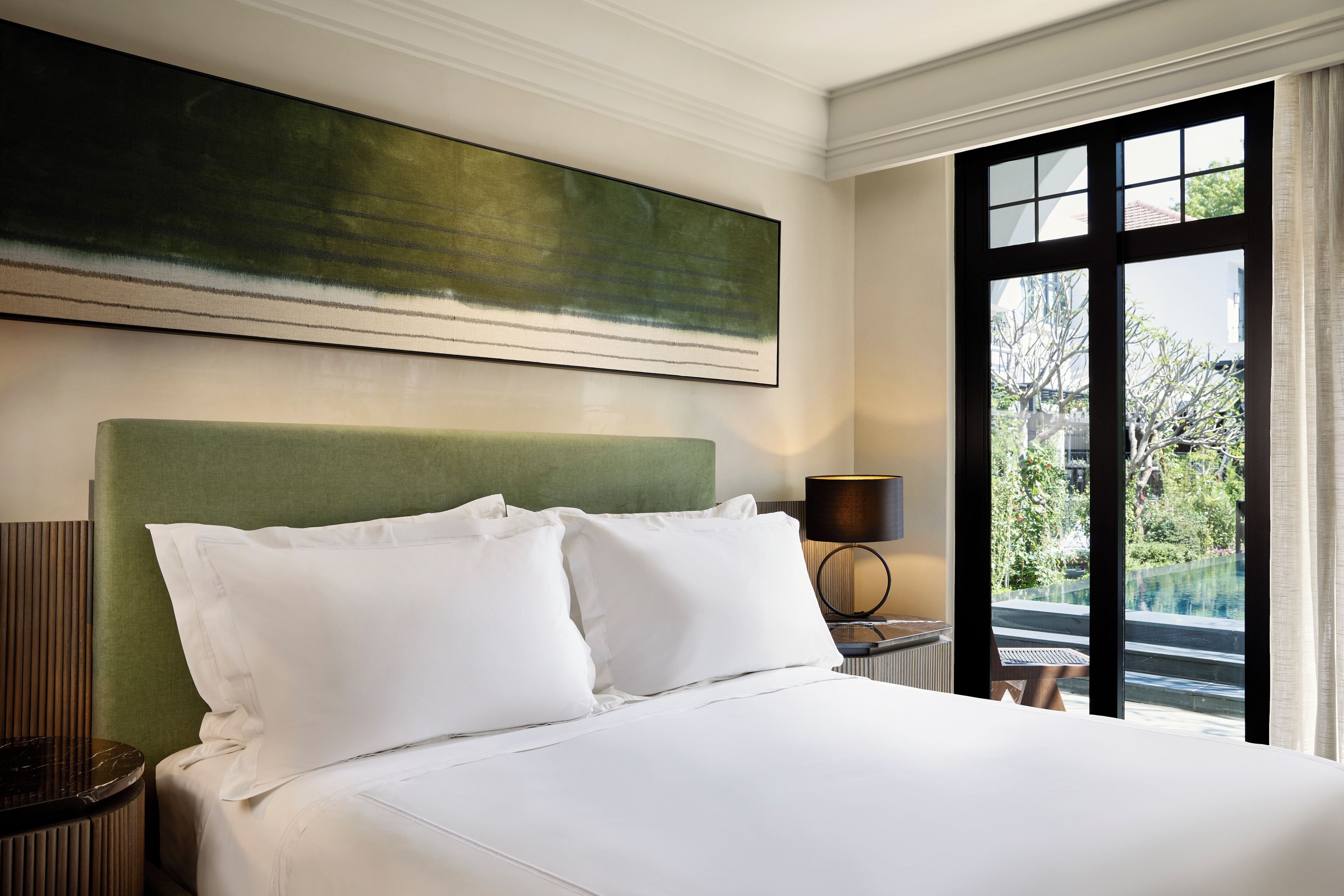 Una habitación de hotel con una cama, cabecera verde, un ventanal grande y un cuadro en la pared.