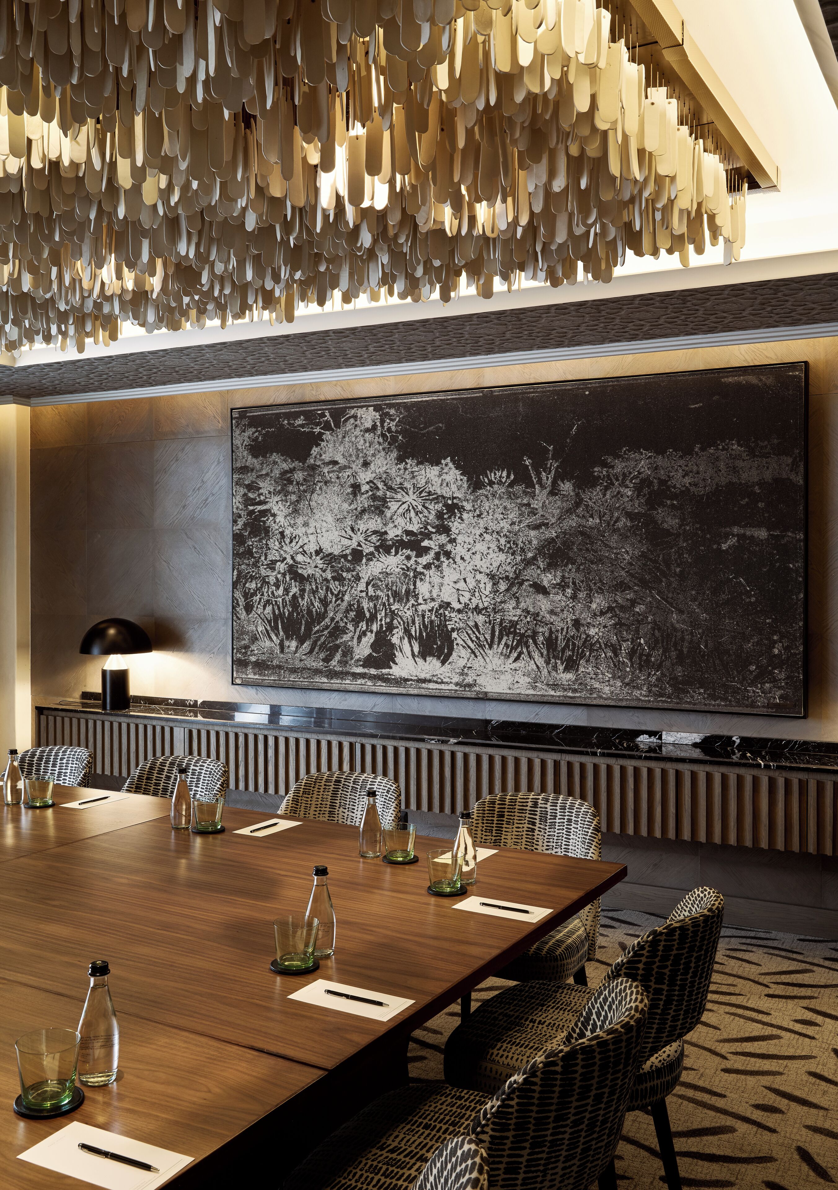 Un salón de conferencias con una gran mesa, sillas de mimbre, un candelabro moderno y una obra de arte grande en blanco y negro en la pared.