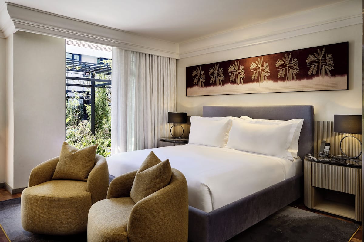 Une chambre d’hôtel avec un grand lit, deux tables de chevet, une vue sur la verdure et une œuvre d’art accrochée au mur.